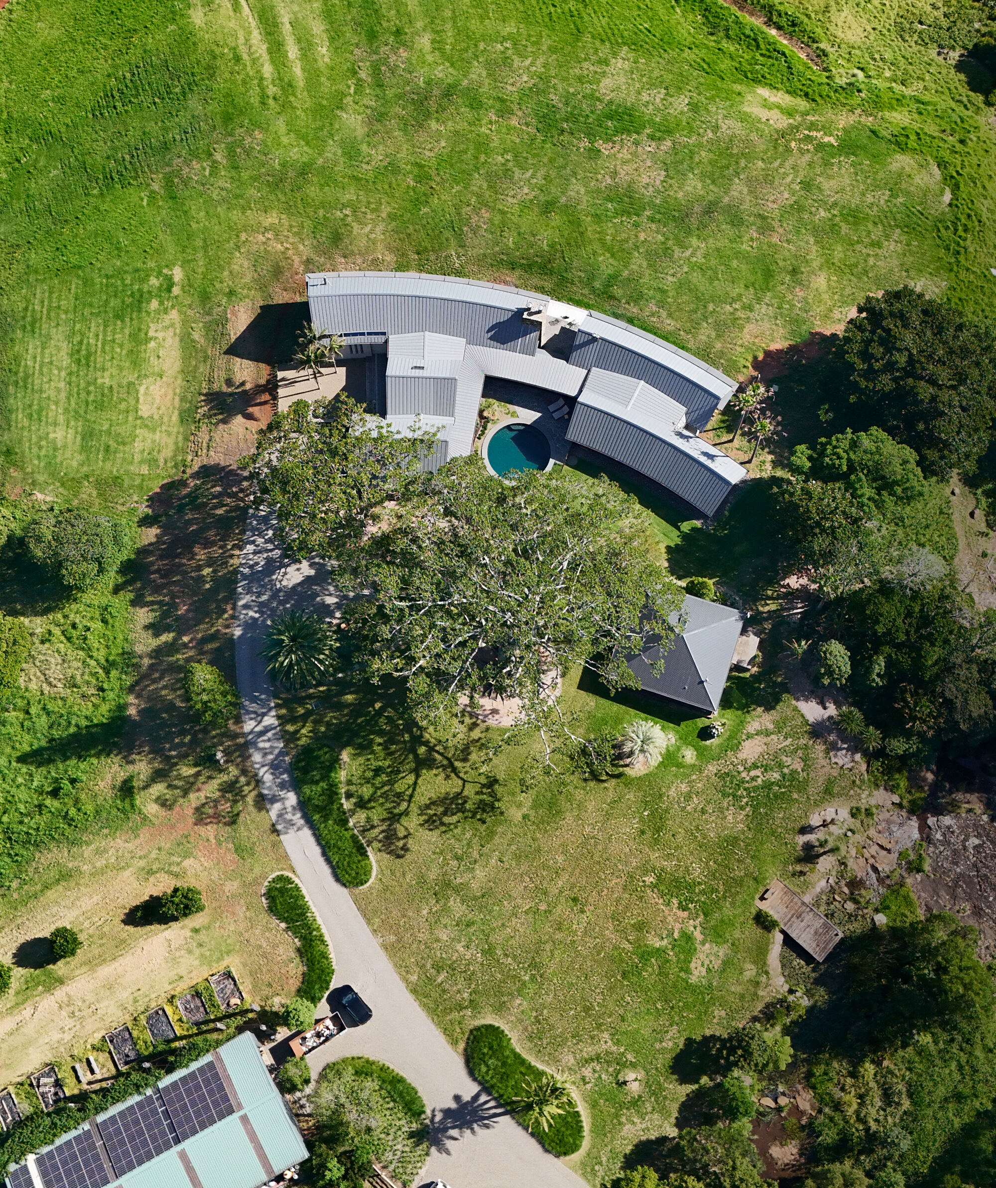 Aerial view showing curved metal roofs encircling a pool within a pastoral landscape of fields and trees