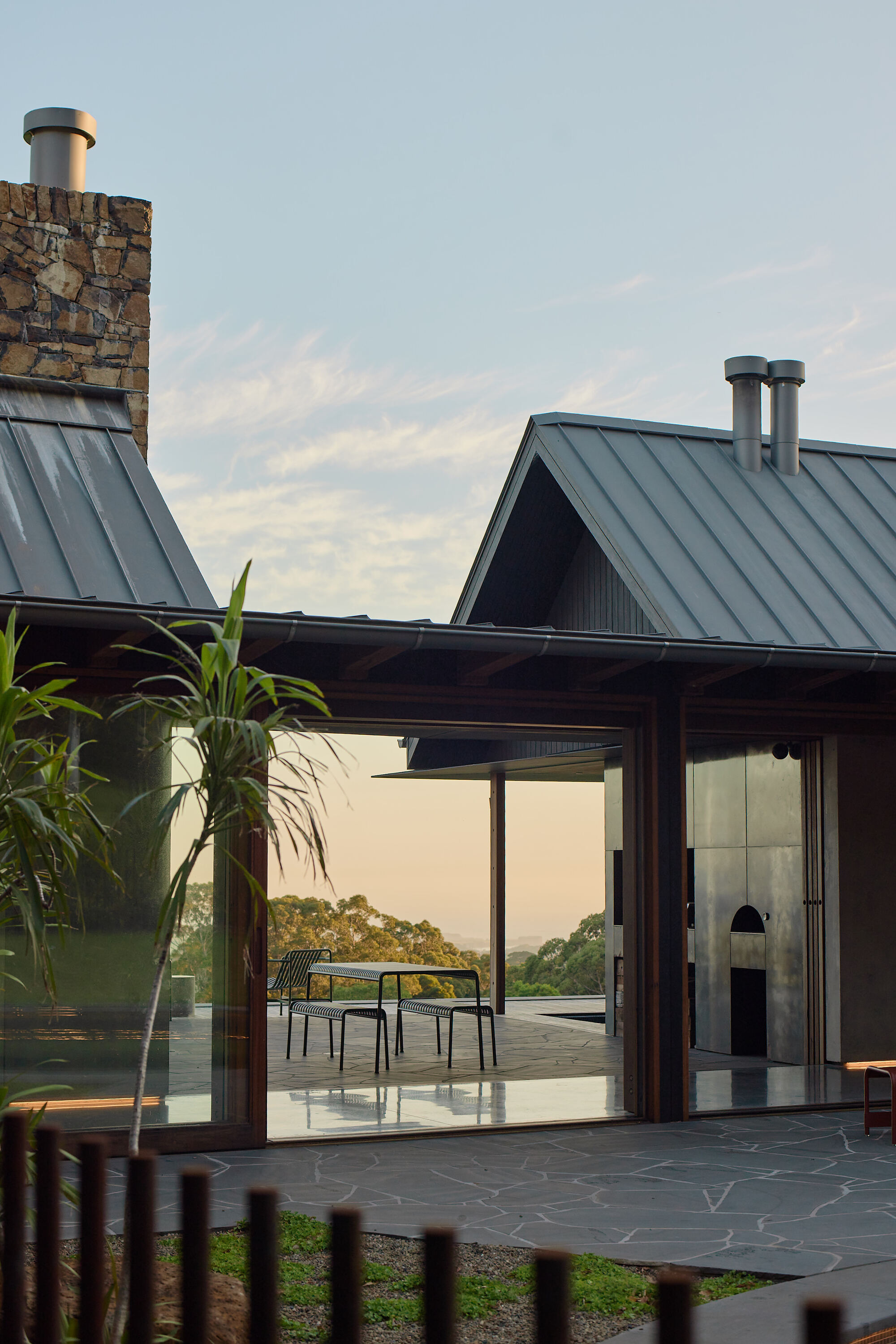 Gabled metal roofs with stone chimney and glazed doorway opening to a terrace at dusk