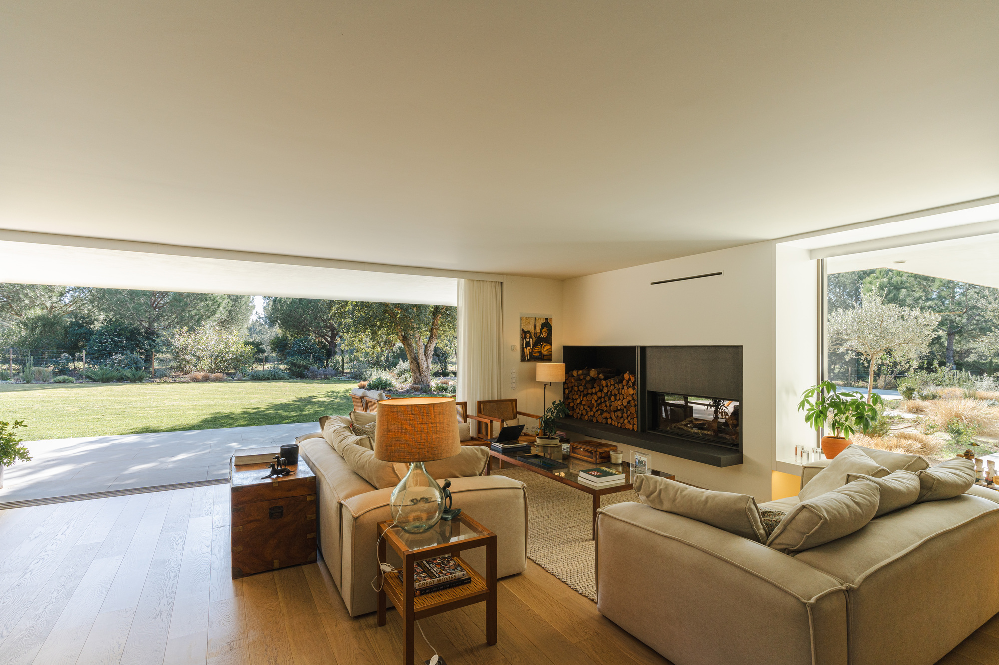 Living room with full-height glazing opening to lawn and fireplace with stacked firewood storage