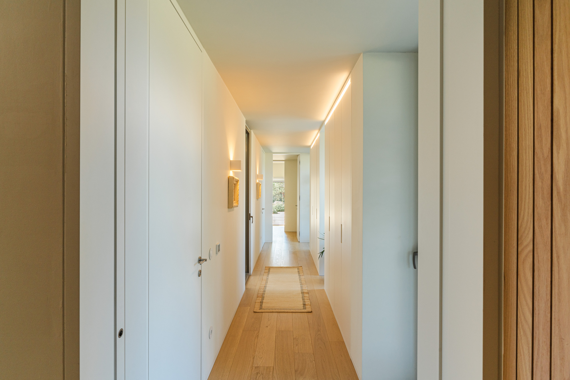 White corridor with timber flooring and concealed wall sconces leading to a distant window