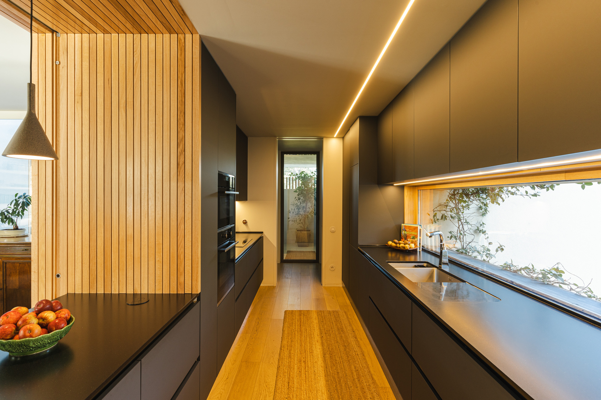 Galley kitchen with timber slat wall and horizontal window overlooking planted courtyard in evening light
