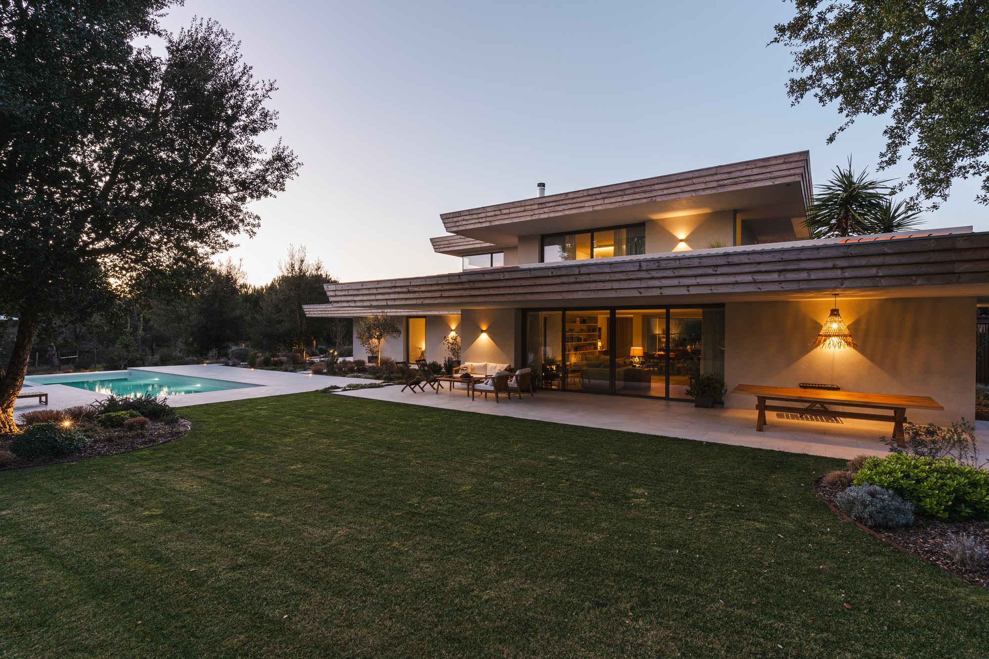 Garden facade with stacked terracotta roof planes and covered terrace overlooking lawn and pool at dusk