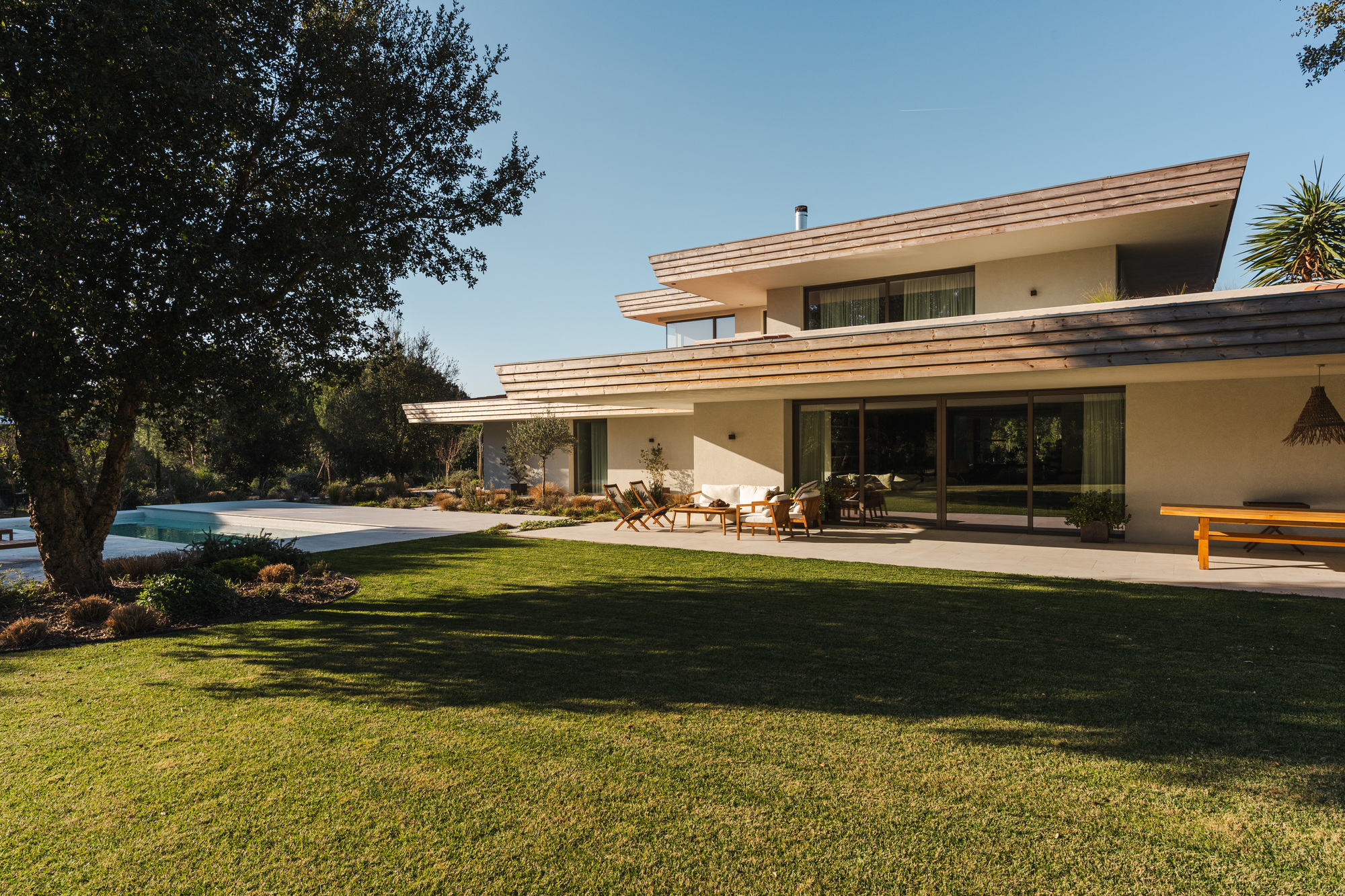 Rear elevation showing layered roof overhangs and glass walls opening to terrace and lawn in morning light