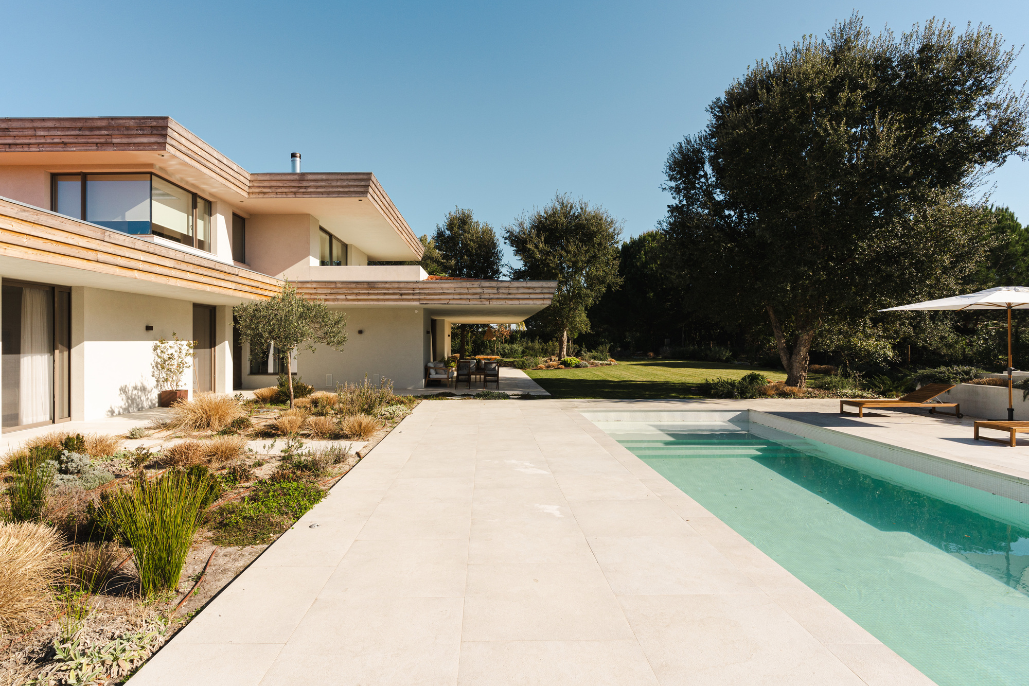 Terrace walkway connecting planted beds to turquoise pool deck under mature olive trees and clear sky