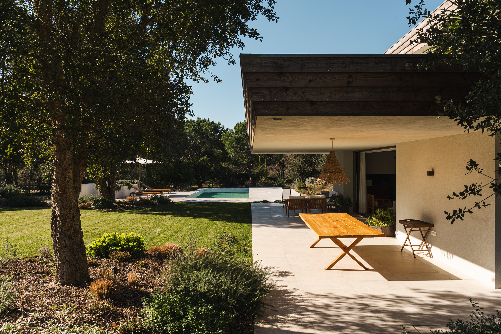 Covered outdoor dining area with timber ceiling extending to garden and pool beneath deciduous trees