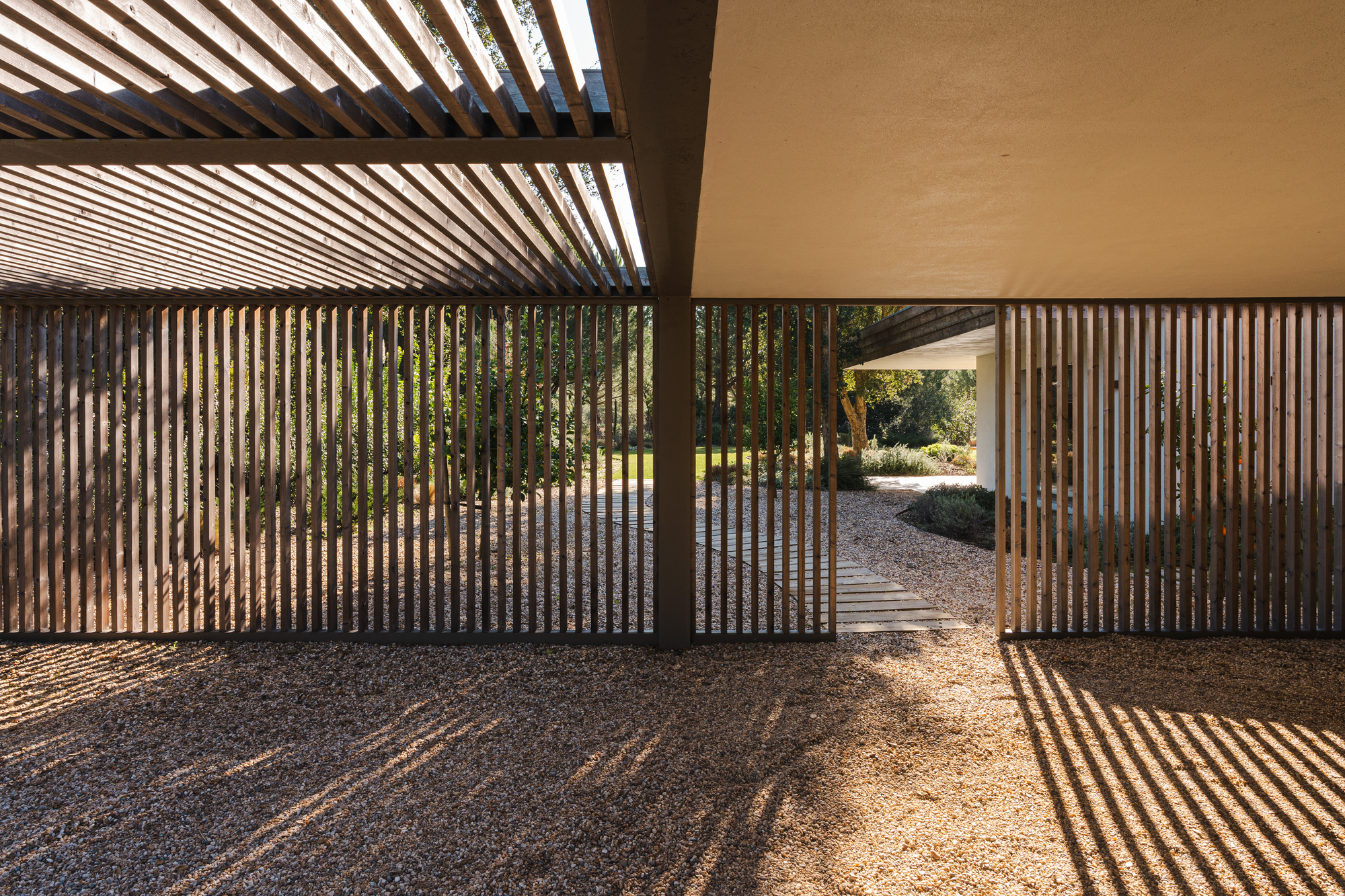 Vertical timber slat screen with pergola overhead casting striped shadows on gravel ground