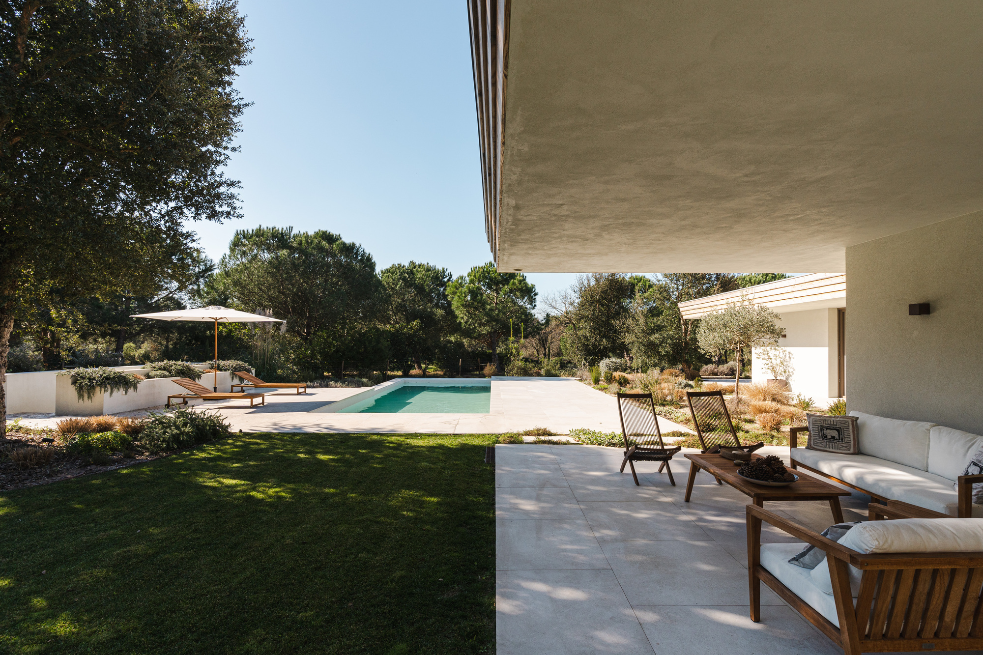 Covered terrace with wooden lounge chairs overlooking the pool and lawn in dappled sunlight