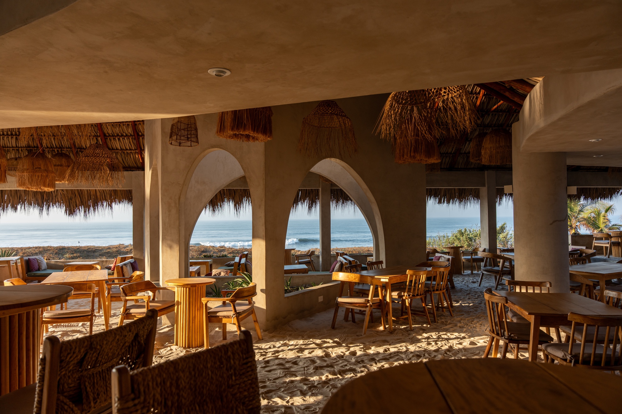 Dining area with earthen arches framing ocean views and dappled sunlight across sand floor
