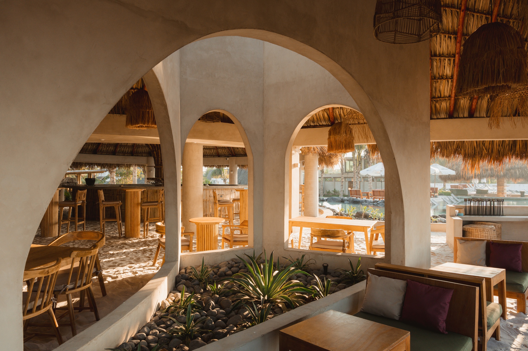 White stucco arches framing a planted bed with agave and a dining area with thatched pendants