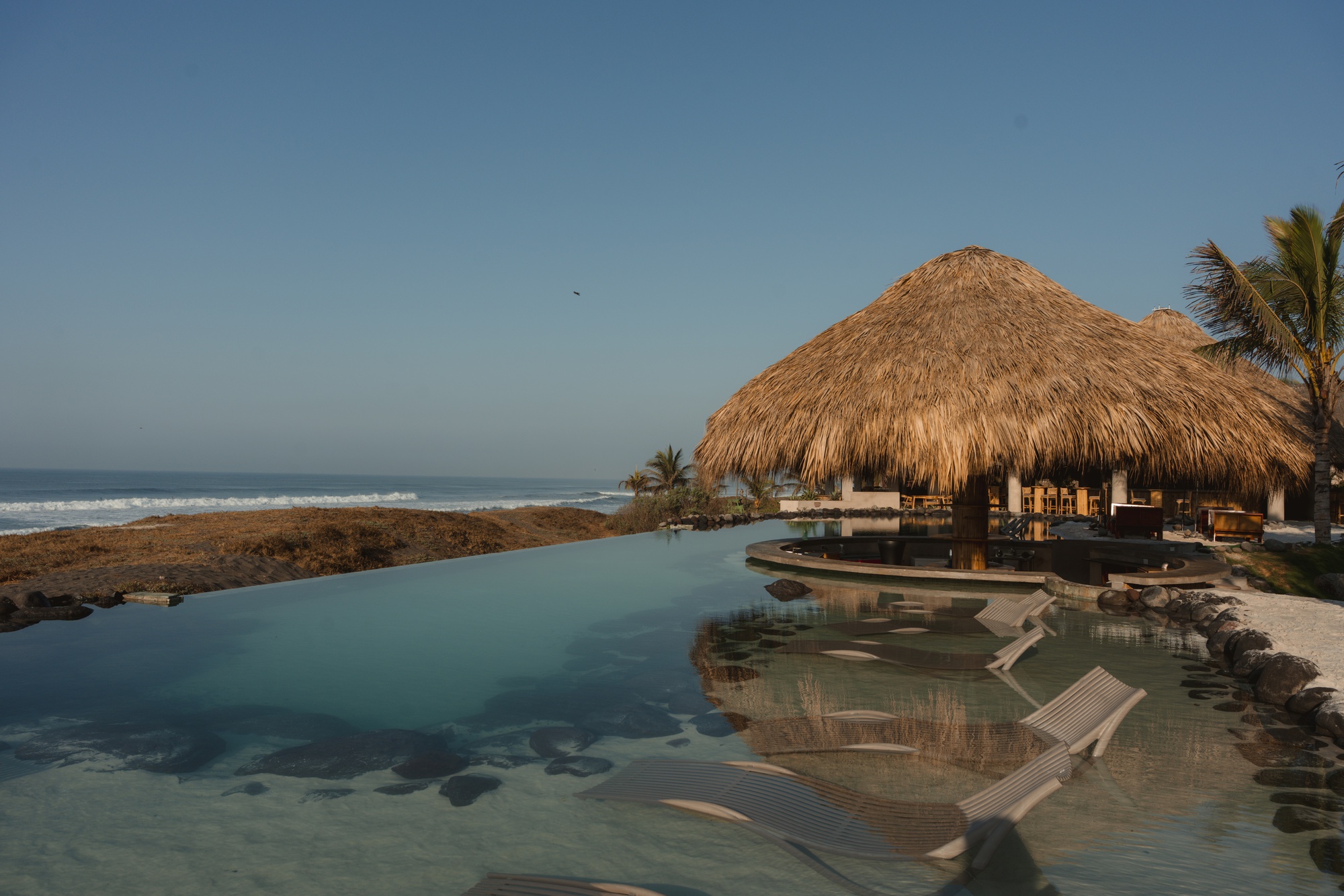 Infinity pool edge with submerged loungers and thatched pavilion against the ocean at dawn