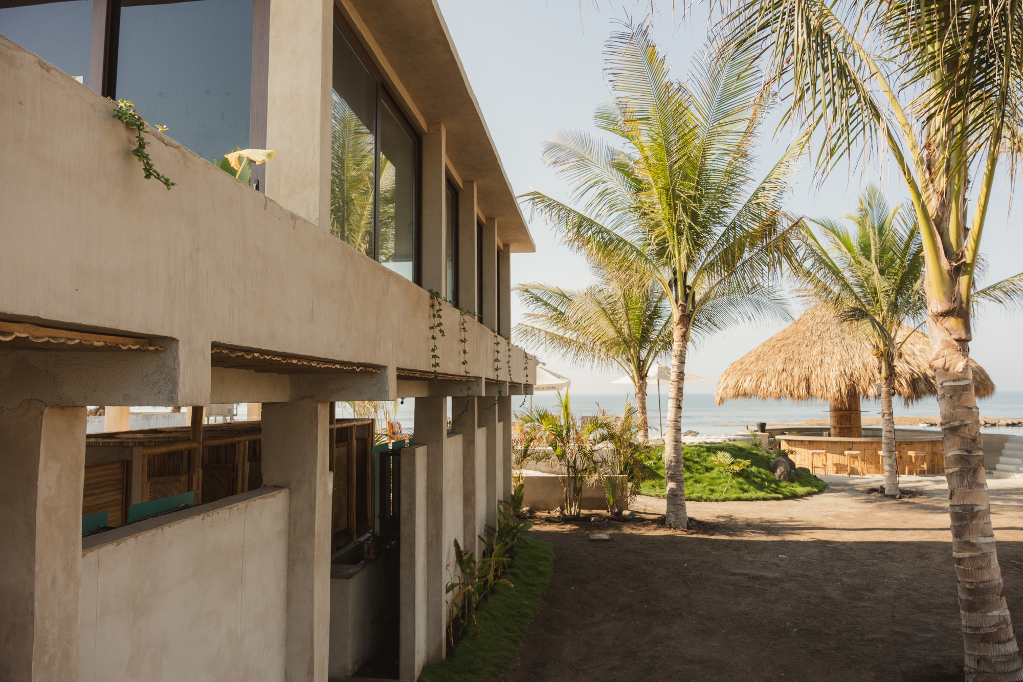 Two-story stucco volume with horizontal windows beside palm trees and thatch-roofed structures near the beach
