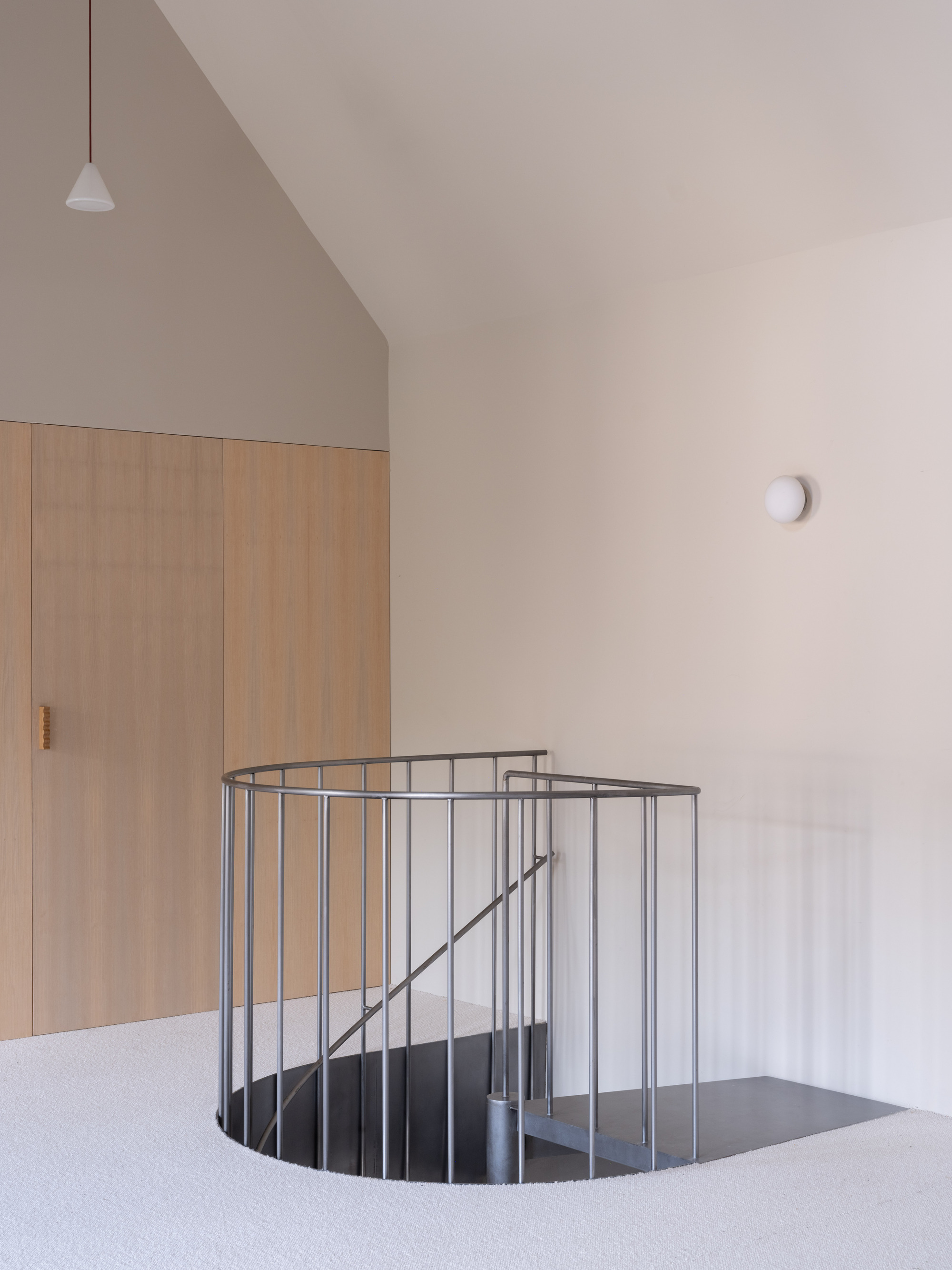 Steel spiral staircase opening framed by plywood wardrobe and white plaster walls under pendant light