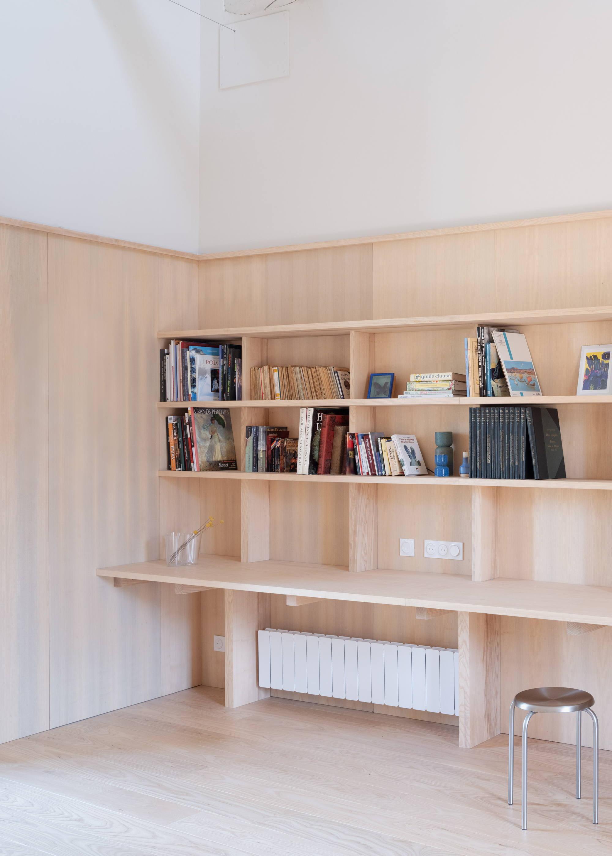Corner built-in pale timber shelving with integrated desk and radiator beneath a white vaulted ceiling