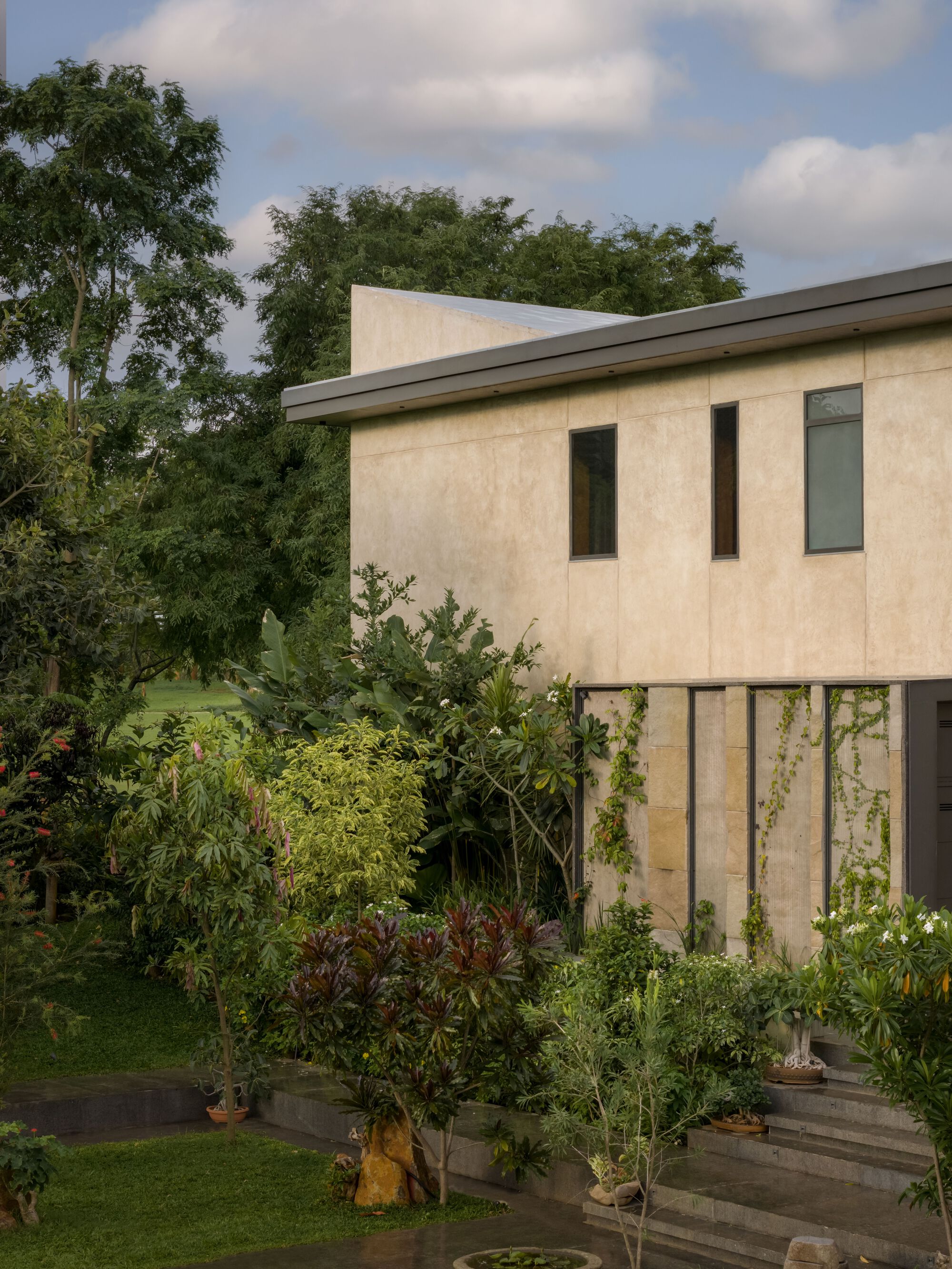 Cream-rendered facade on pilotis with narrow vertical windows and lush garden planting below