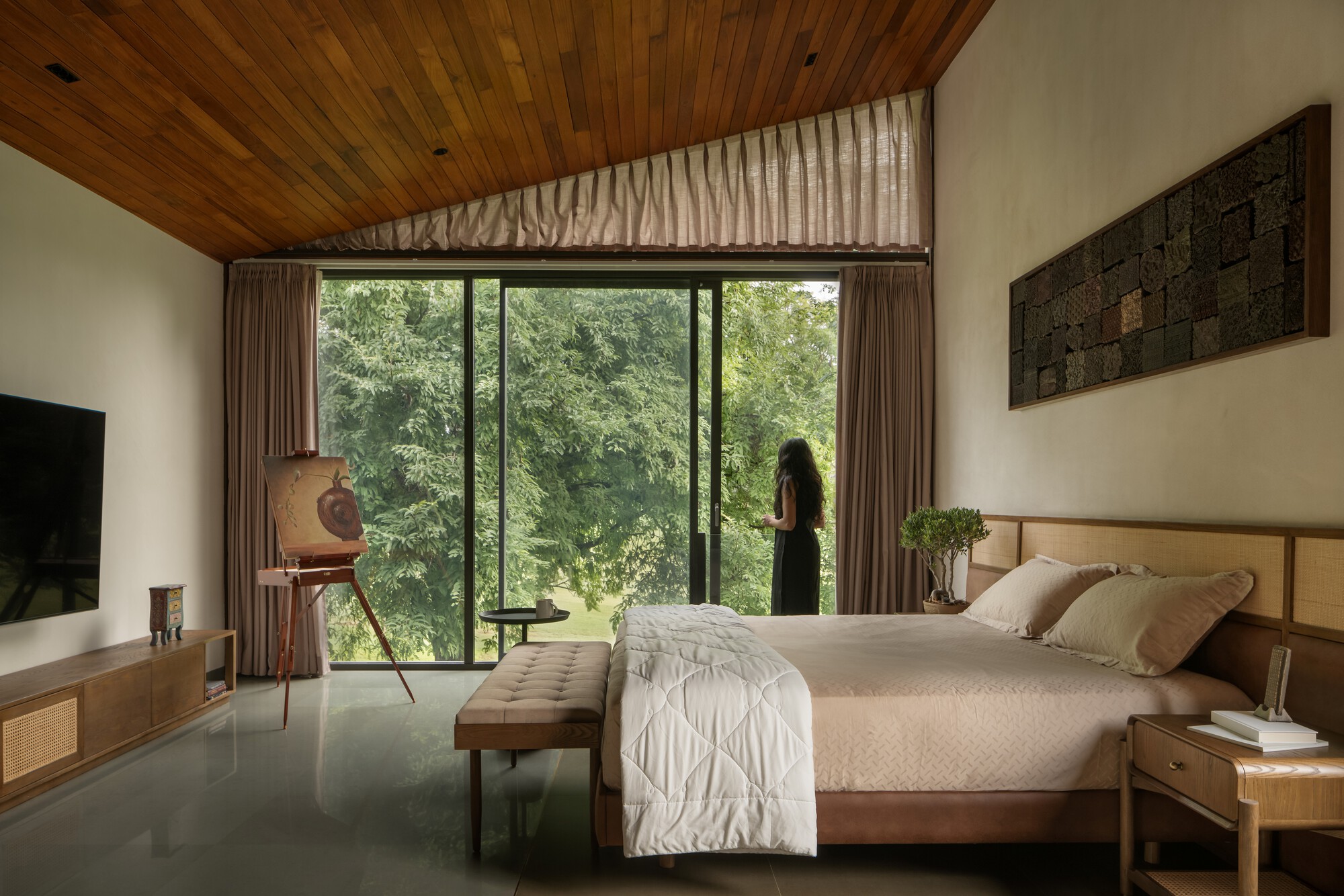 Bedroom with timber ceiling, floor-to-ceiling window framing forest view and figure silhouetted against glass