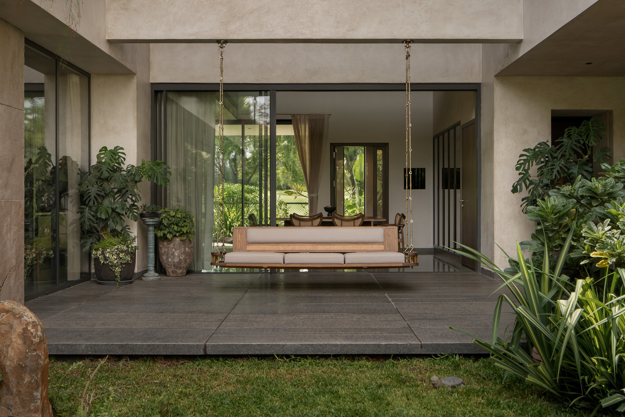 Covered terrace with suspended wooden swing bench opening to planted courtyard through floor-to-ceiling glazing