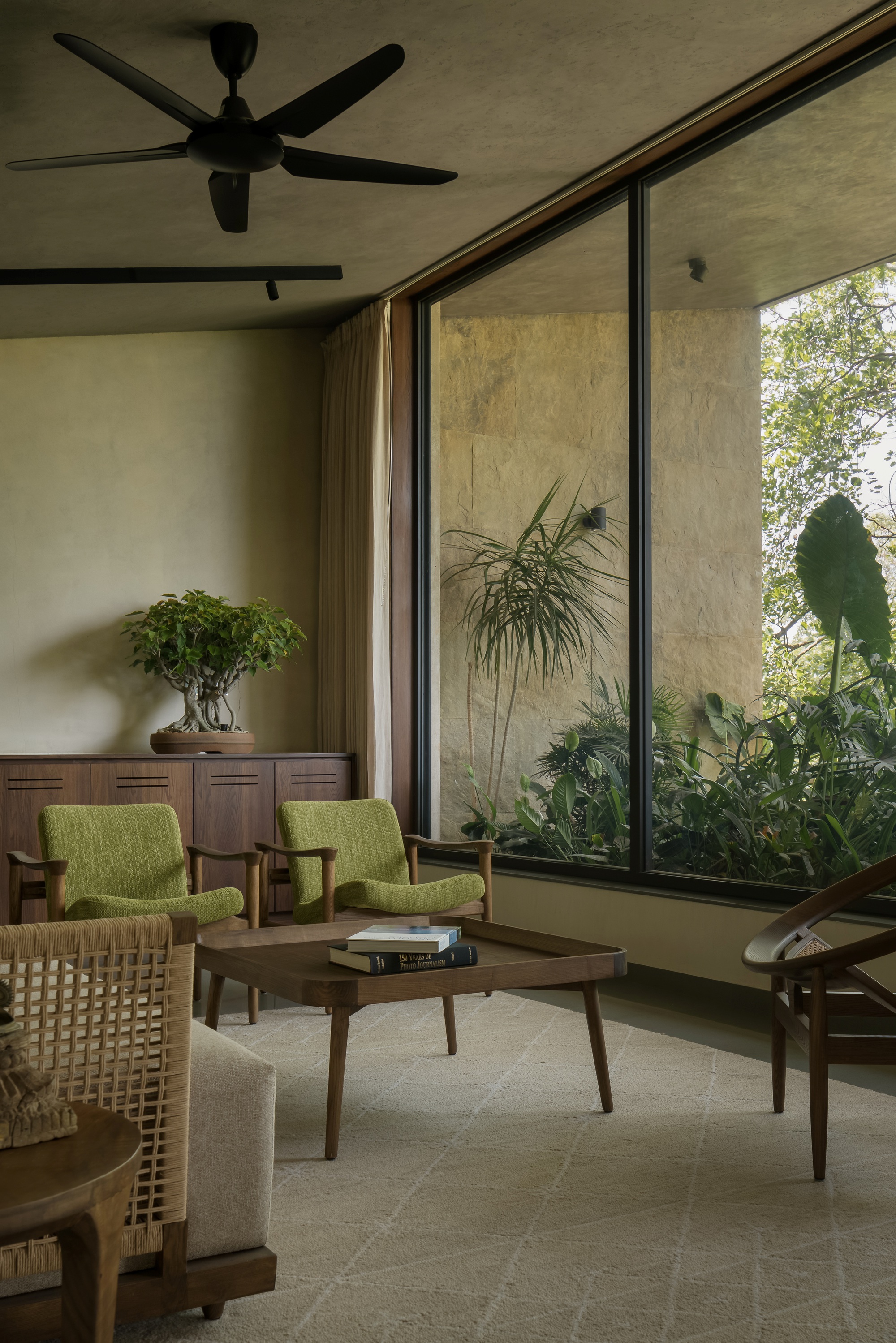 Living room corner with full-height glazing overlooking planted courtyard and ceiling fan above upholstered seating