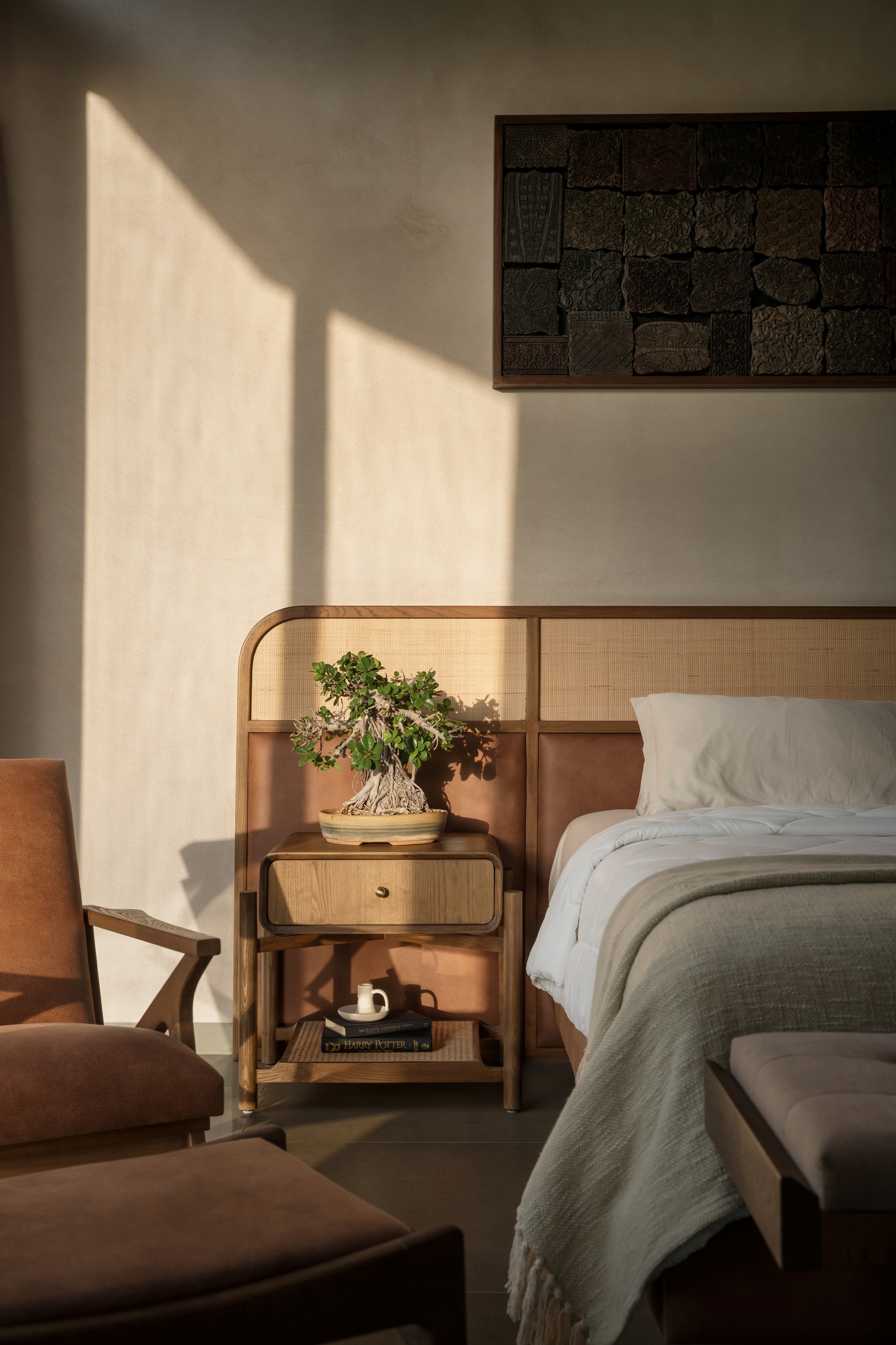 Bedroom nightstand in warm afternoon sunlight with bonsai tree and cane-panel detailing below artwork