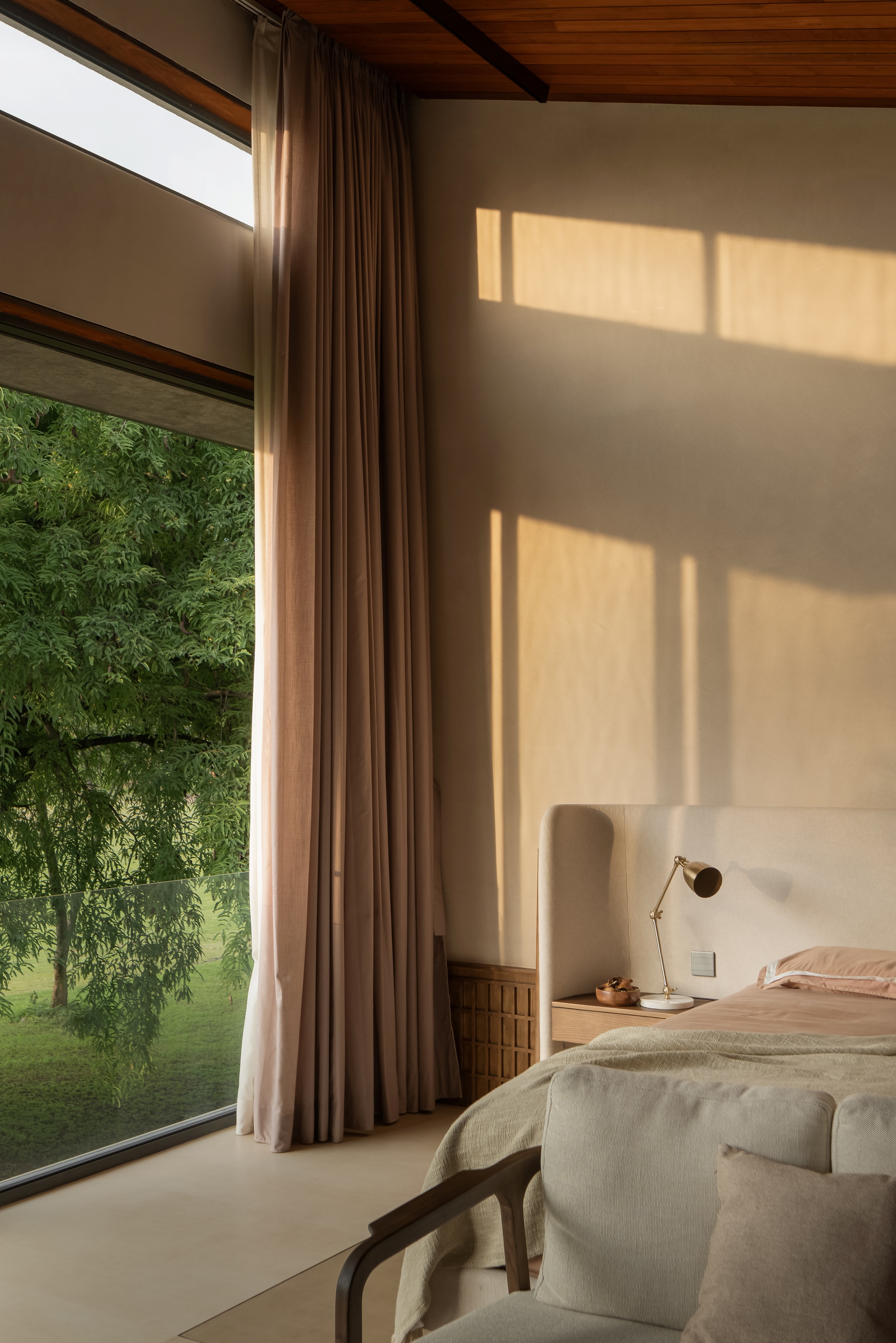 Bedroom with full-height glazing overlooking garden trees, warm sunlight casting rectangular shadows on wall