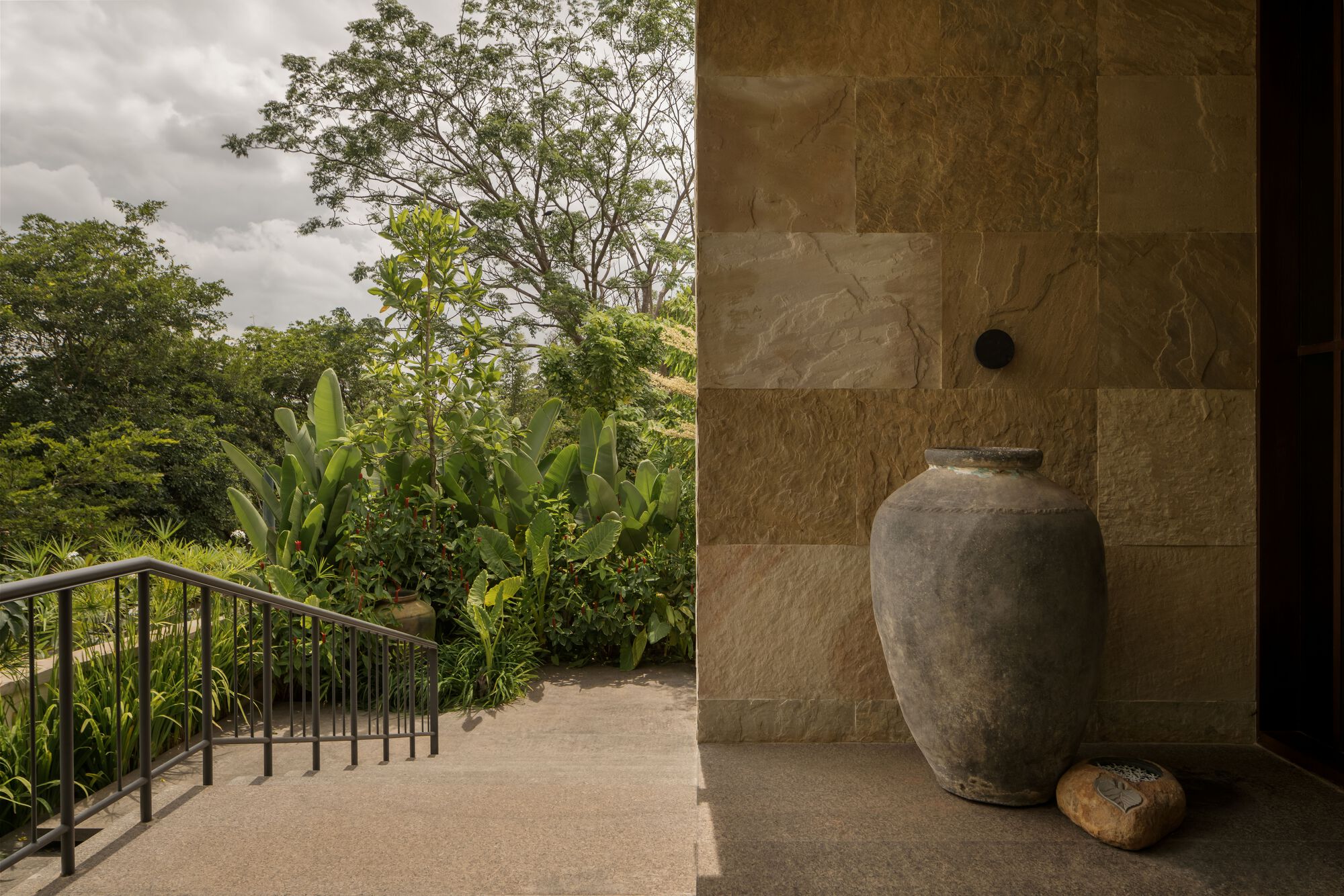 Entry threshold with stone-clad wall, ceramic urn, and metal railing leading to planted hillside