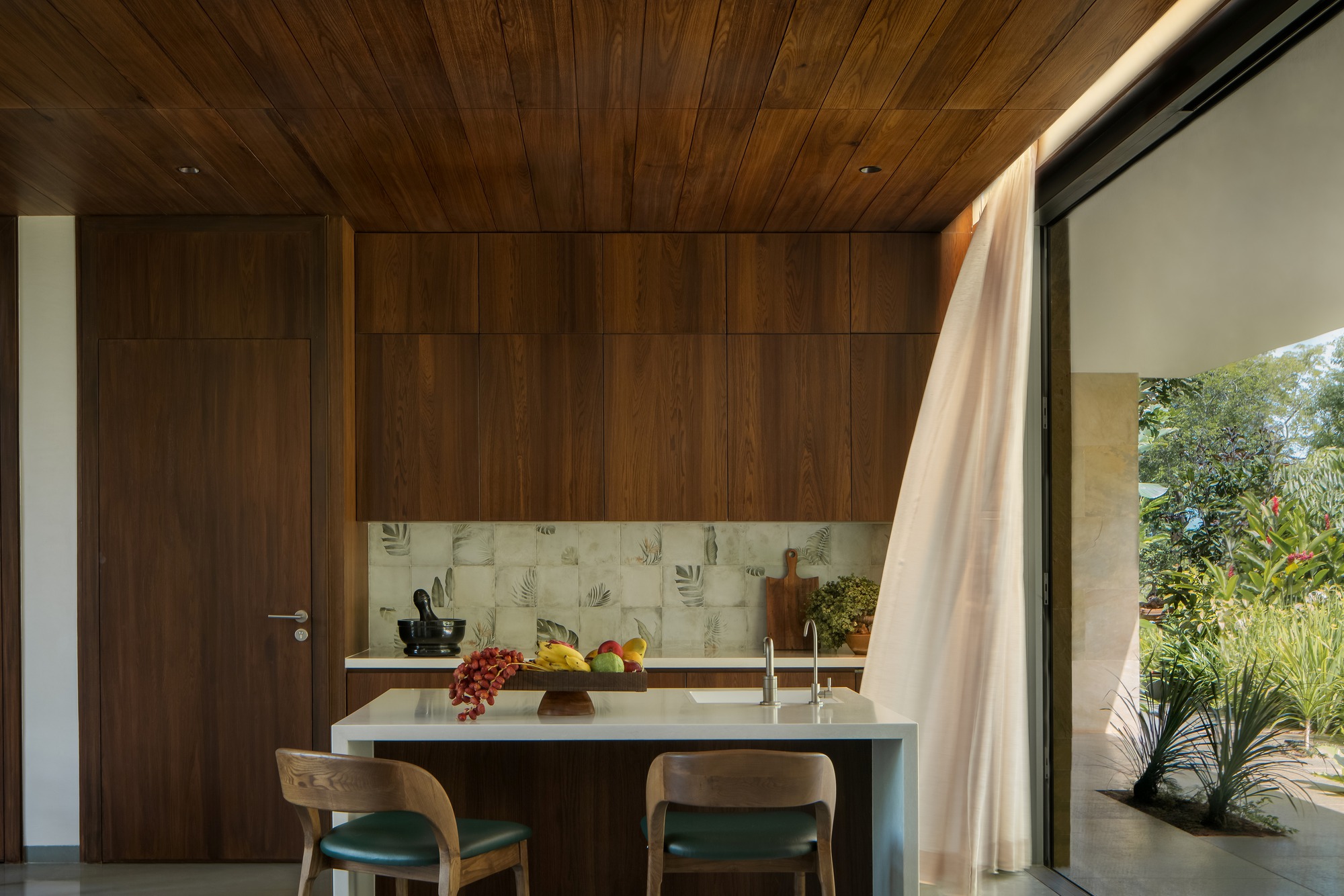Kitchen with walnut cabinetry, patterned tile backsplash, and island opening to planted courtyard