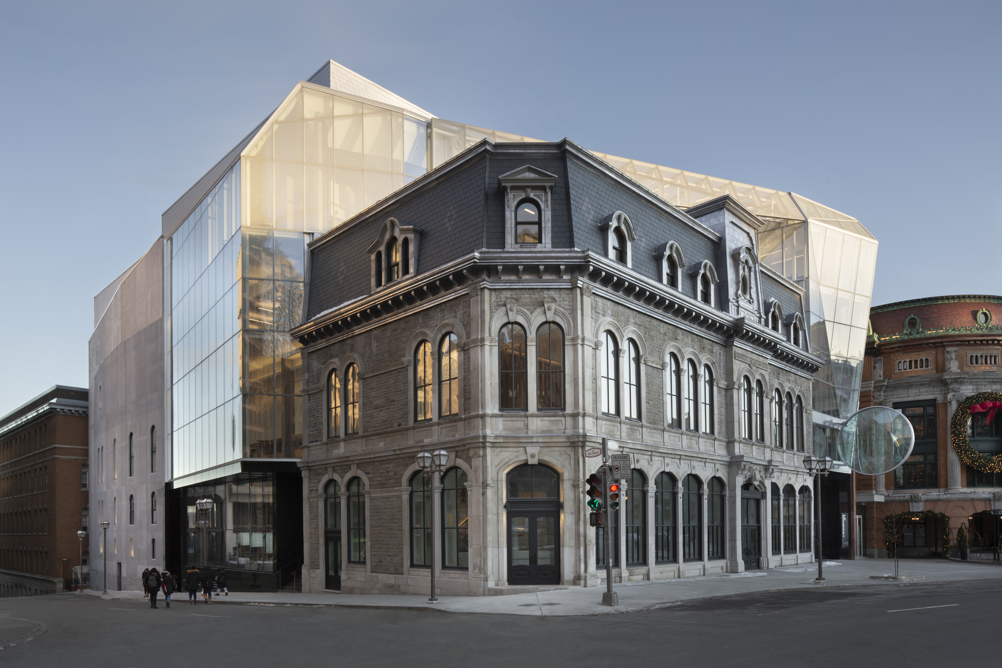 Street corner view of the historic masonry facade wrapped by a translucent glass volume at dusk