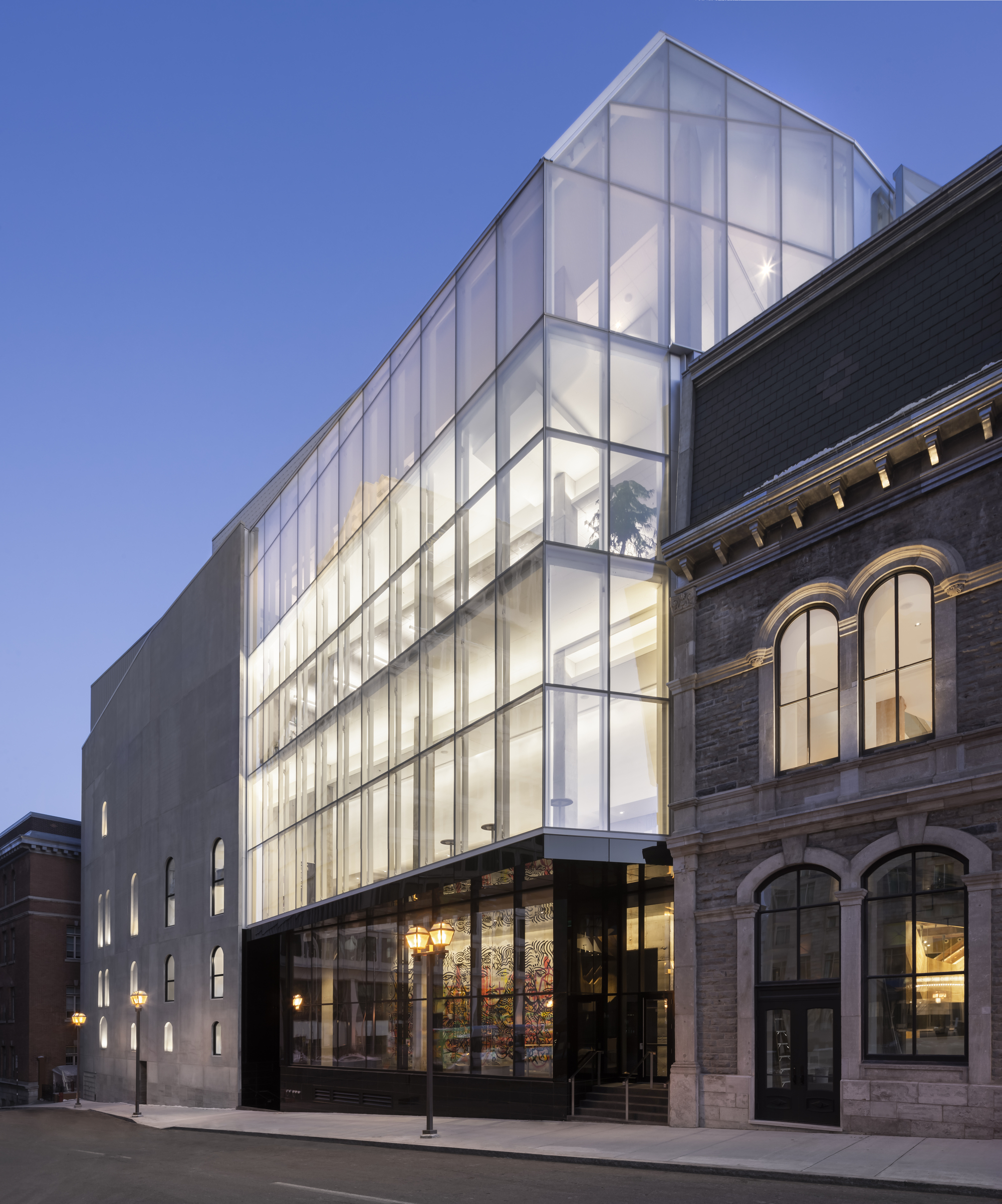 Side elevation showing the illuminated translucent glass facade rising behind the historic brick facade at dusk