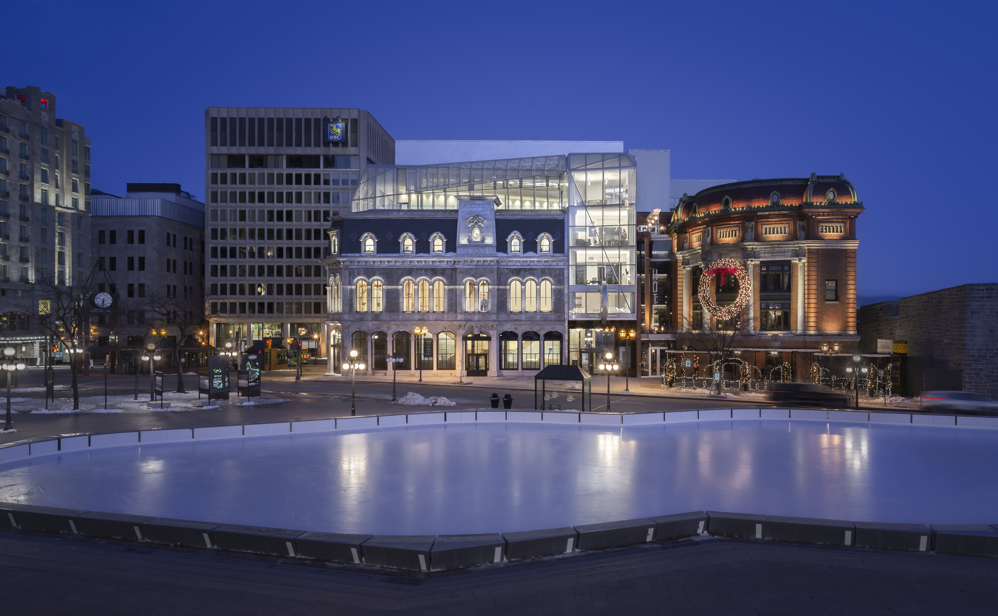 Street view of illuminated glass and concrete addition rising above masonry facades at dusk