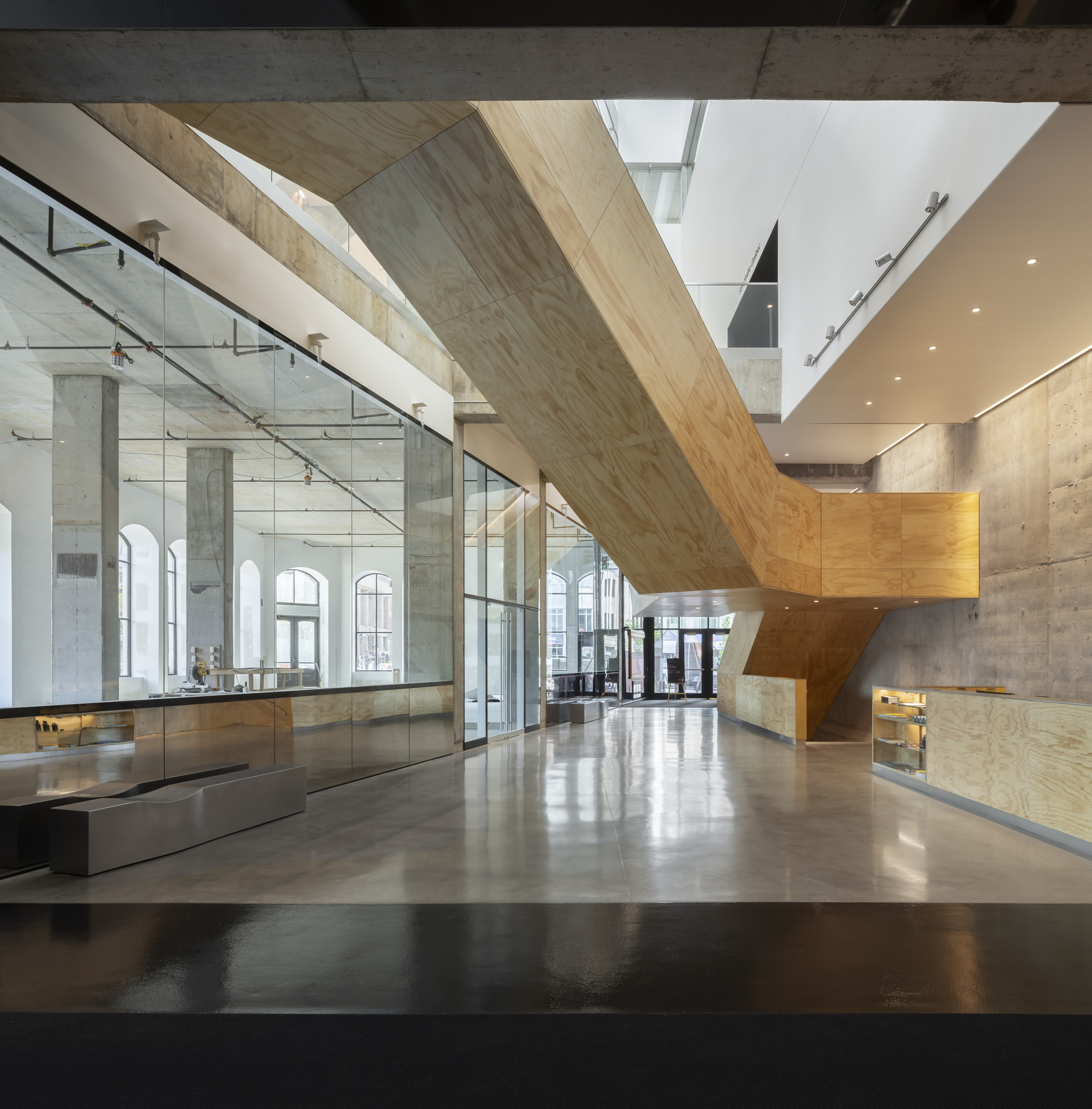Entrance lobby with angular plywood-clad staircase beside glass-enclosed gallery and polished concrete floor