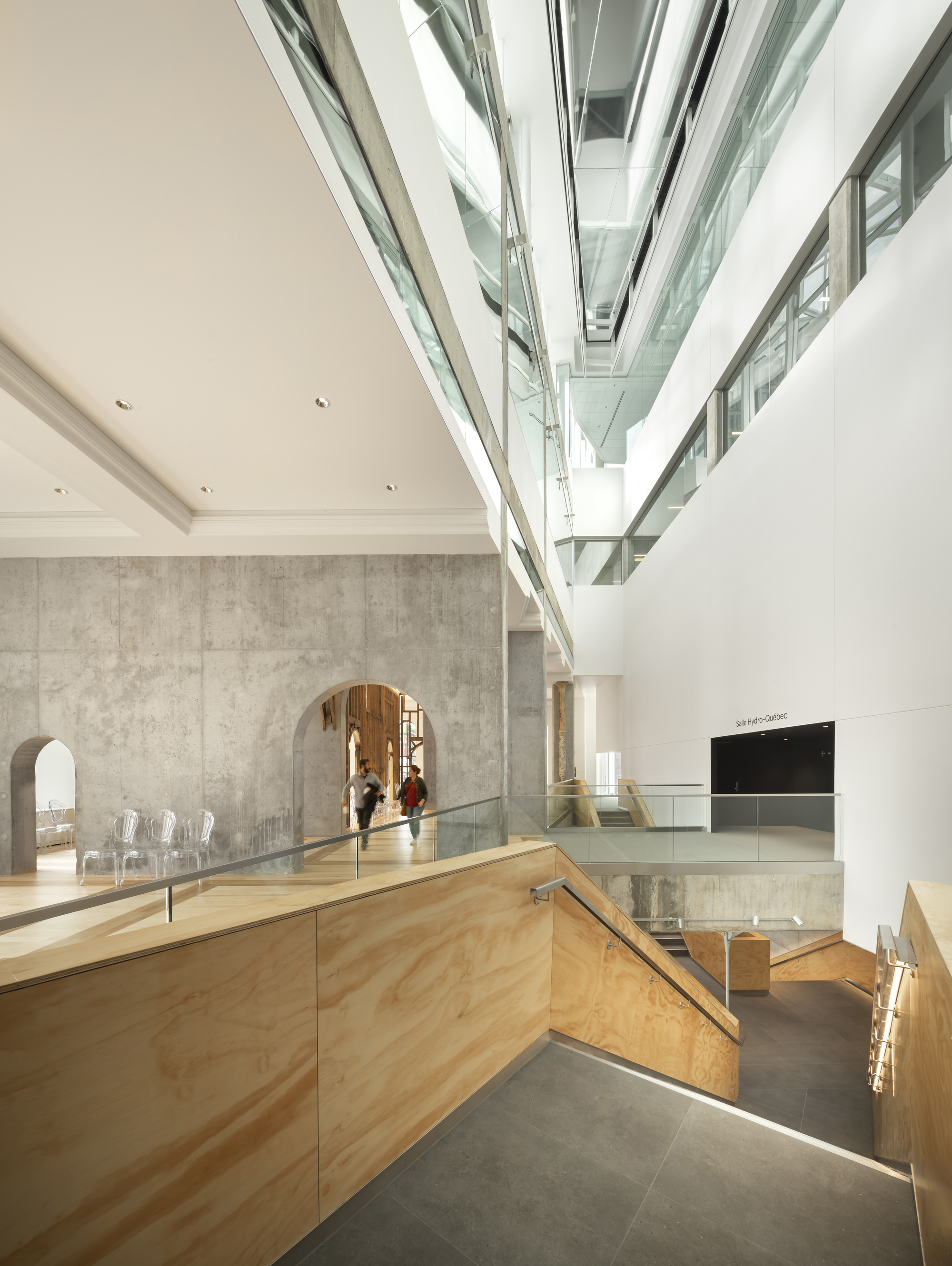 Multi-story atrium with glass balustrades, concrete walls with arched openings, and oak flooring under natural daylight