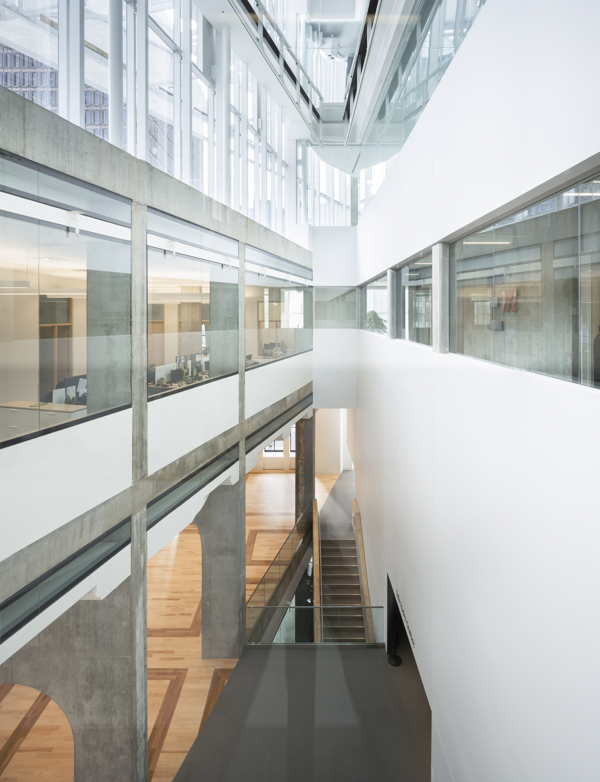 Interior atrium showing concrete beams, glass-enclosed corridors, and a staircase connecting multiple levels under an overcast sky