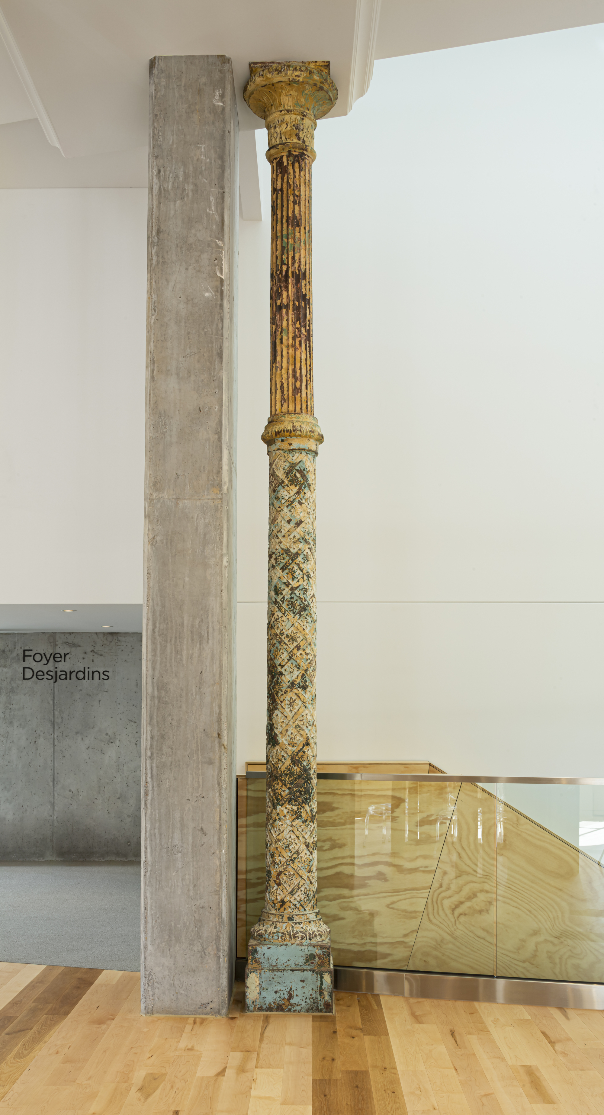 Gilded historic column standing beside a concrete beam and glass balustrade on polished timber flooring