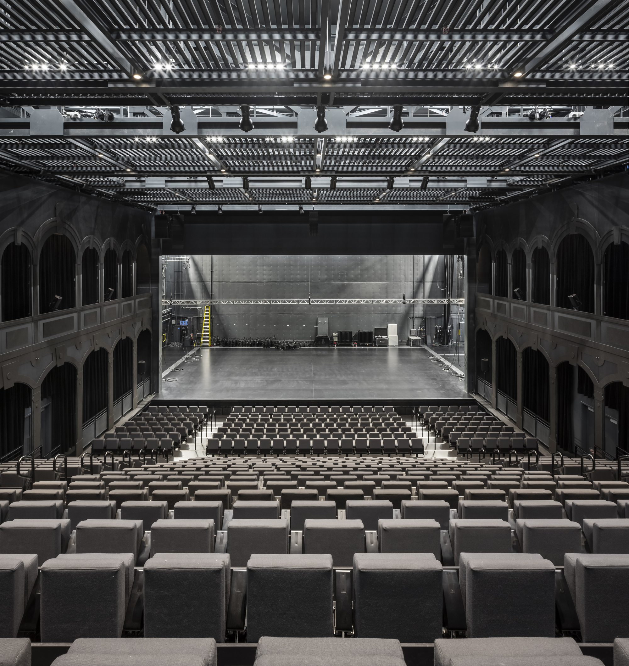 Theatre auditorium with rows of seating facing a steel fly tower beneath exposed technical grid ceiling