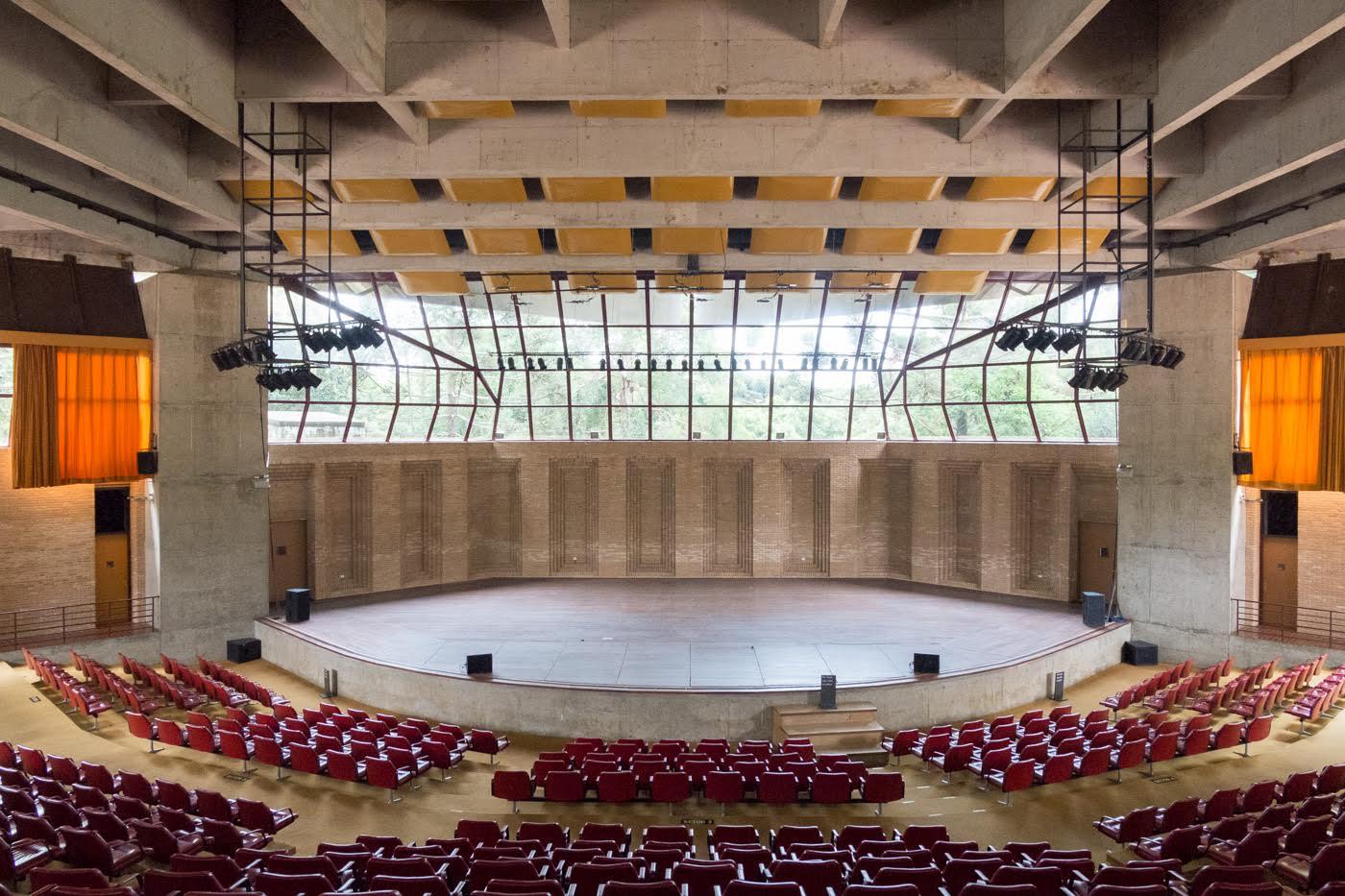 Concert hall interior with exposed concrete ceiling, curved stage backdrop, red seating, and glazed rear wall