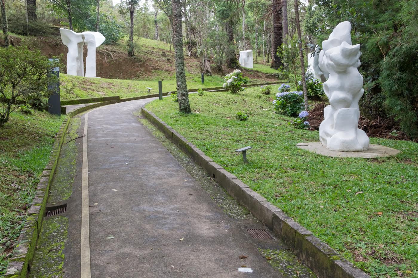 Concrete walkway with drainage channels winding through a sculpture garden with white figures and trees