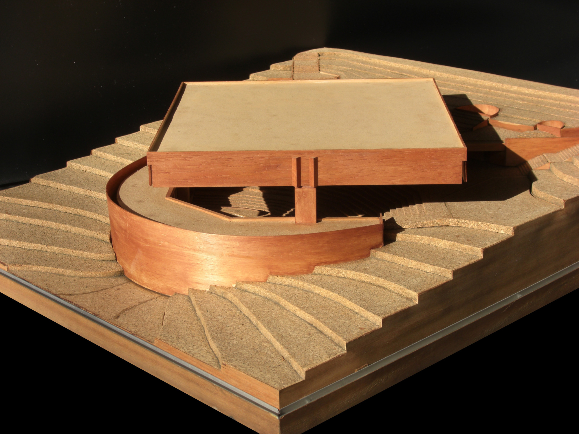 Physical model showing a curved wooden volume and rectangular pavilion on a terraced hillside