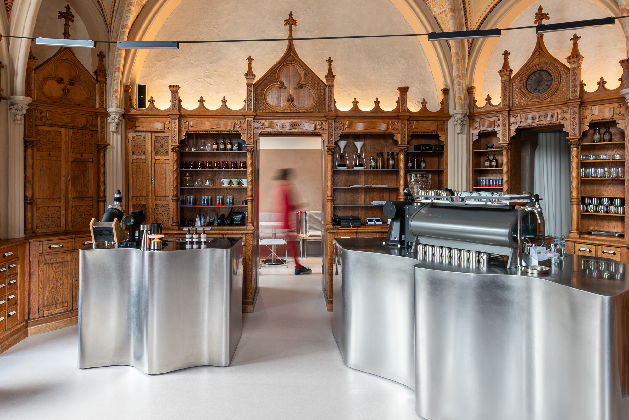 Sculptural stainless steel counters beneath carved wooden cabinetry with Gothic spires and vaulted ceiling