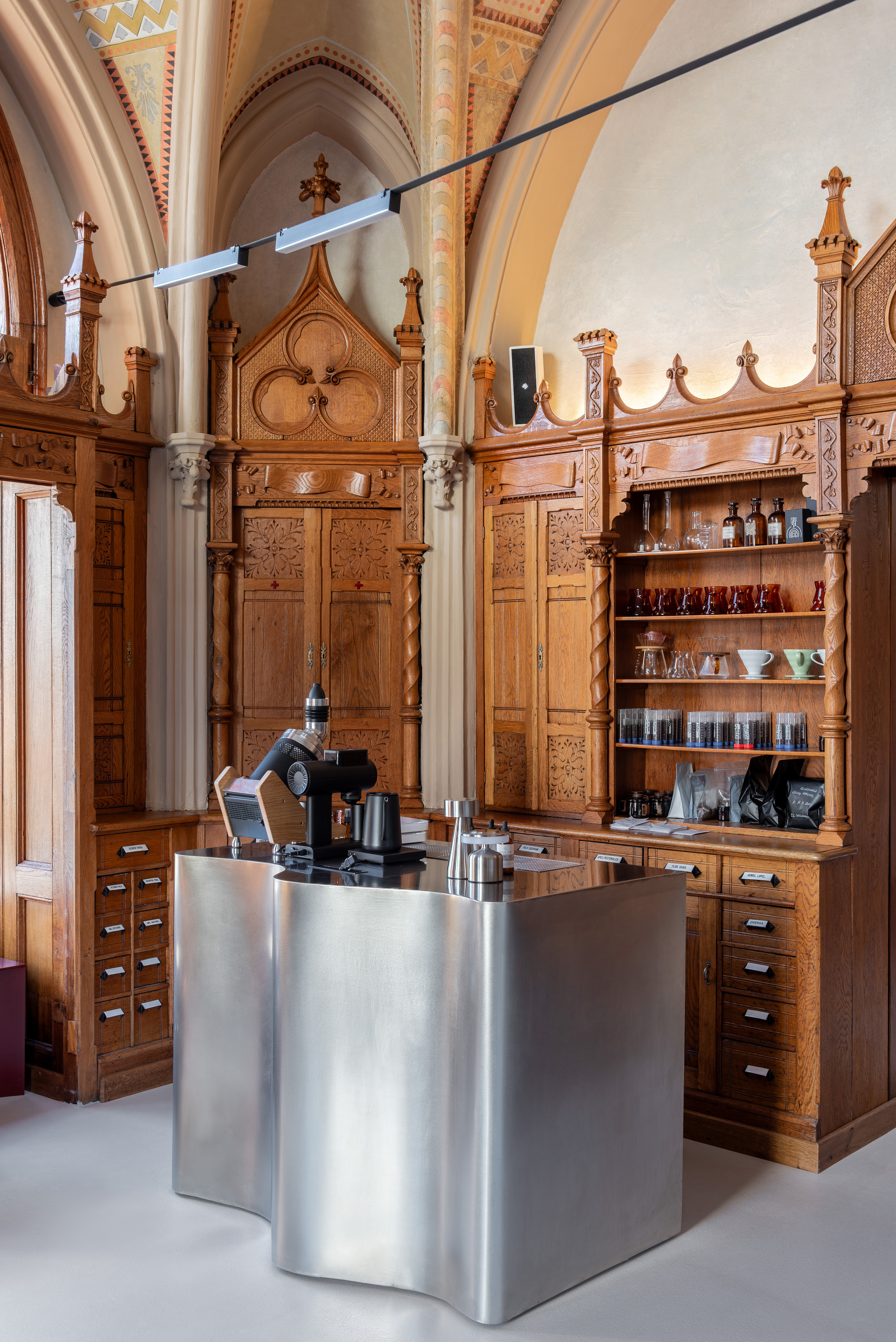Single undulating stainless steel counter with coffee equipment below pointed arch cabinetry and painted vaults