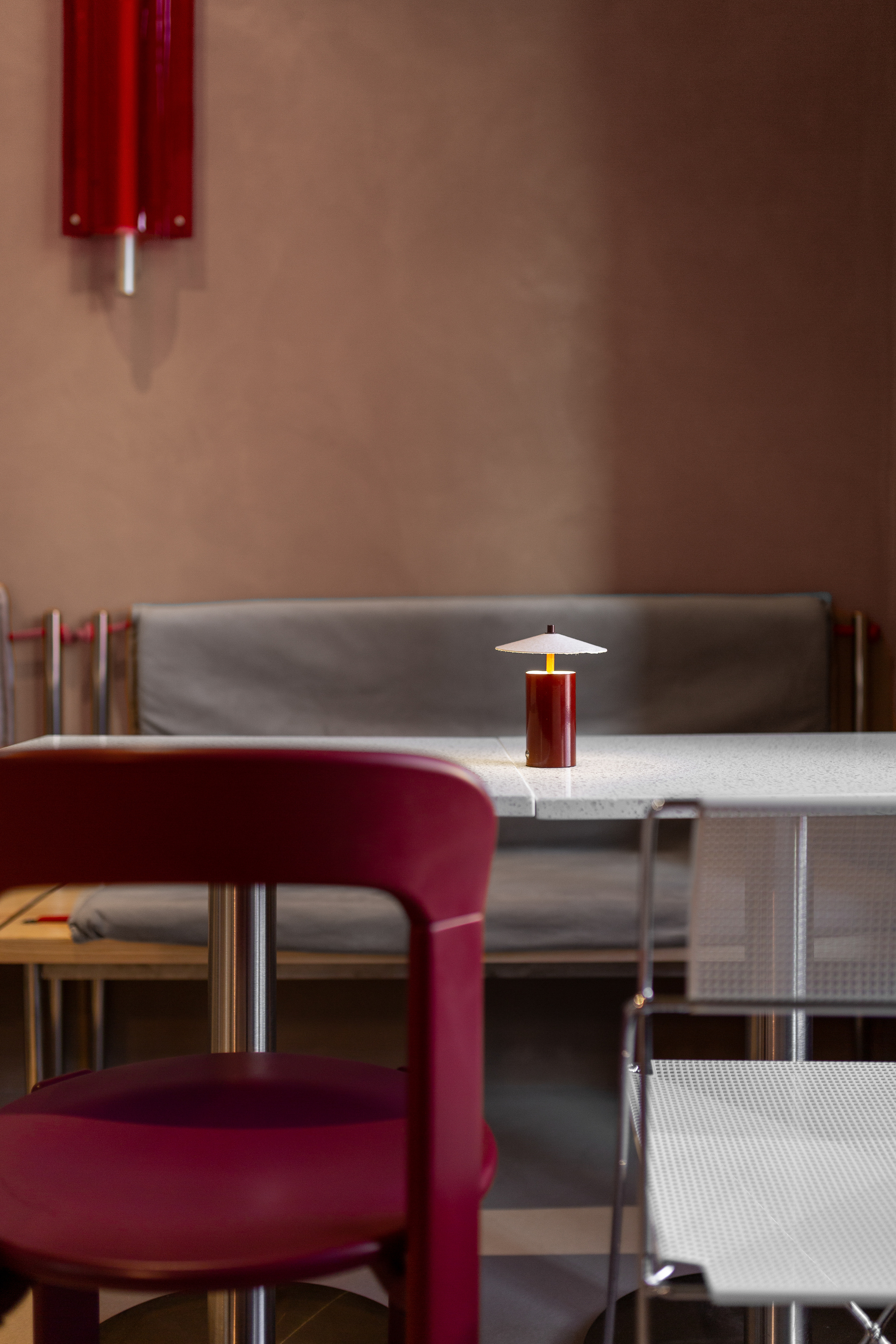 Maroon upholstered chair beside white table with red cylindrical pendant lamp in soft light
