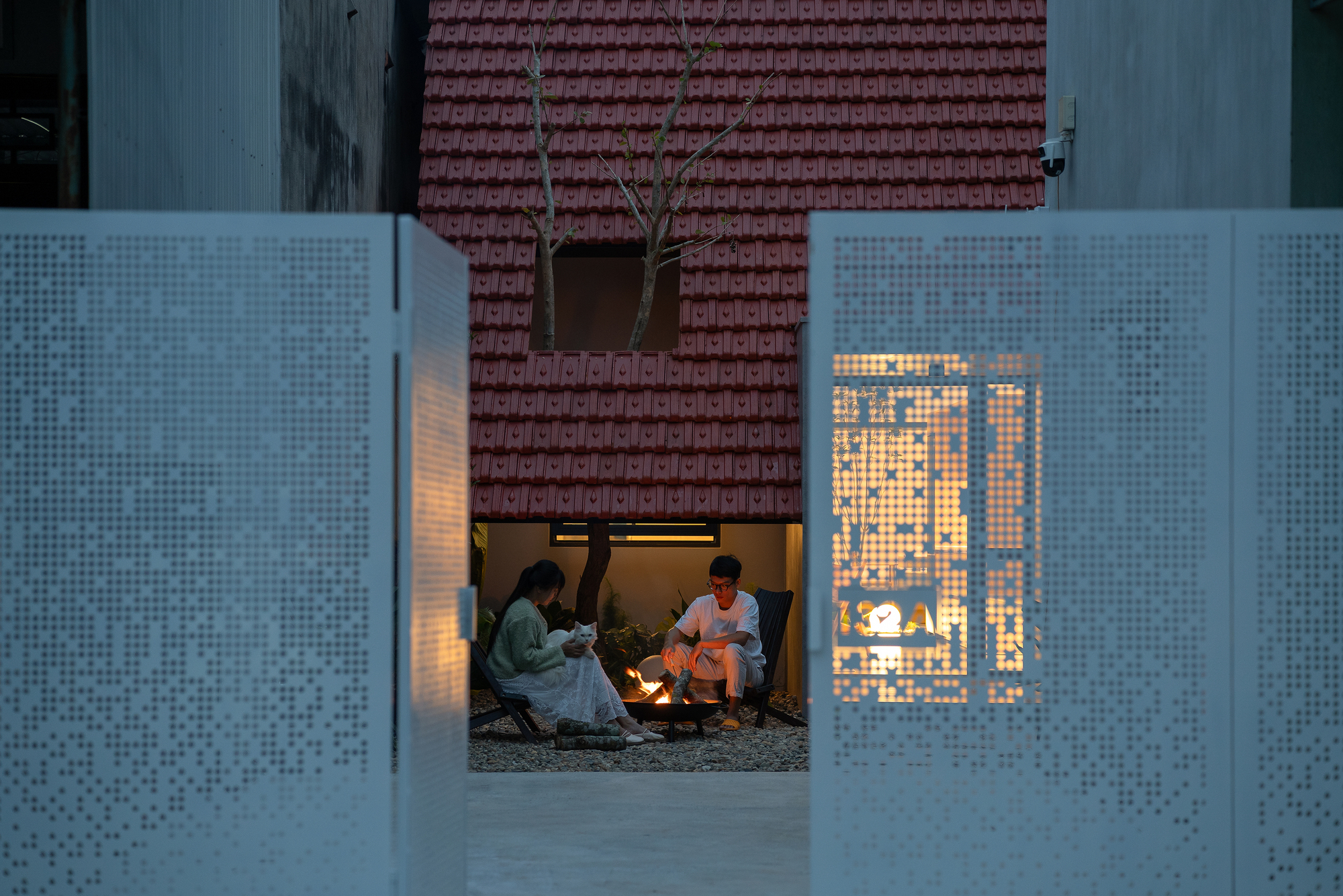 View through perforated white metal screens into a courtyard where two people sit at dusk