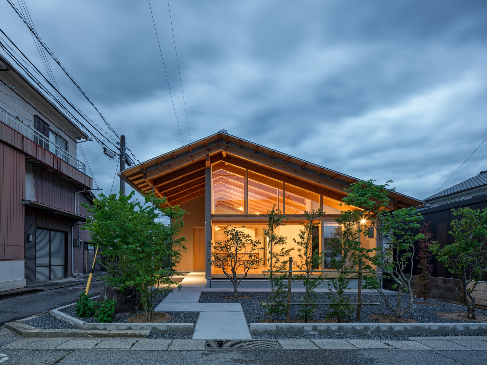 Evening view of the illuminated gable facade with exposed rafters and planted forecourt at dusk