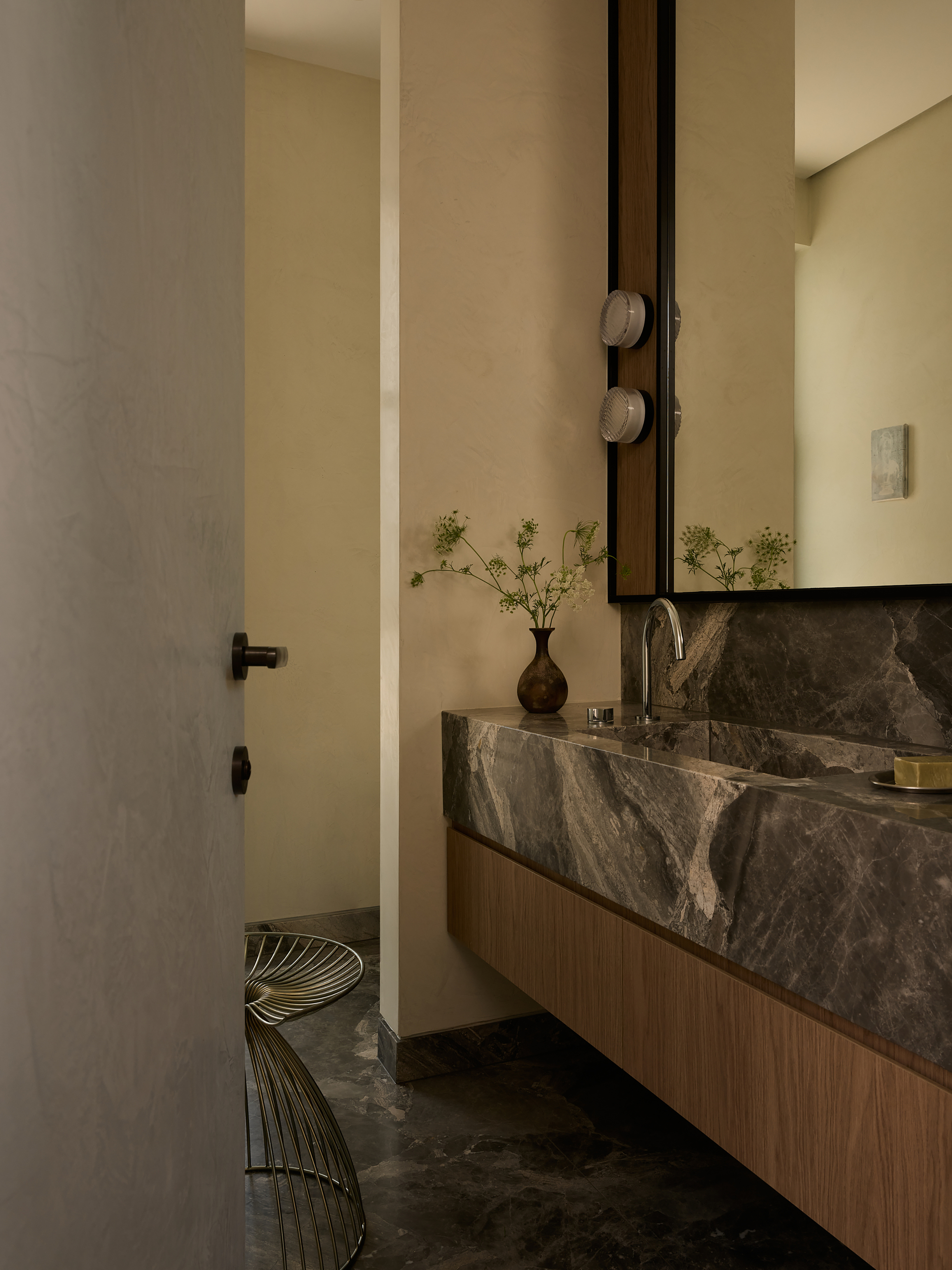 Bathroom with dark stone floating vanity, vertical mirror, and wire stool on stone tile floor