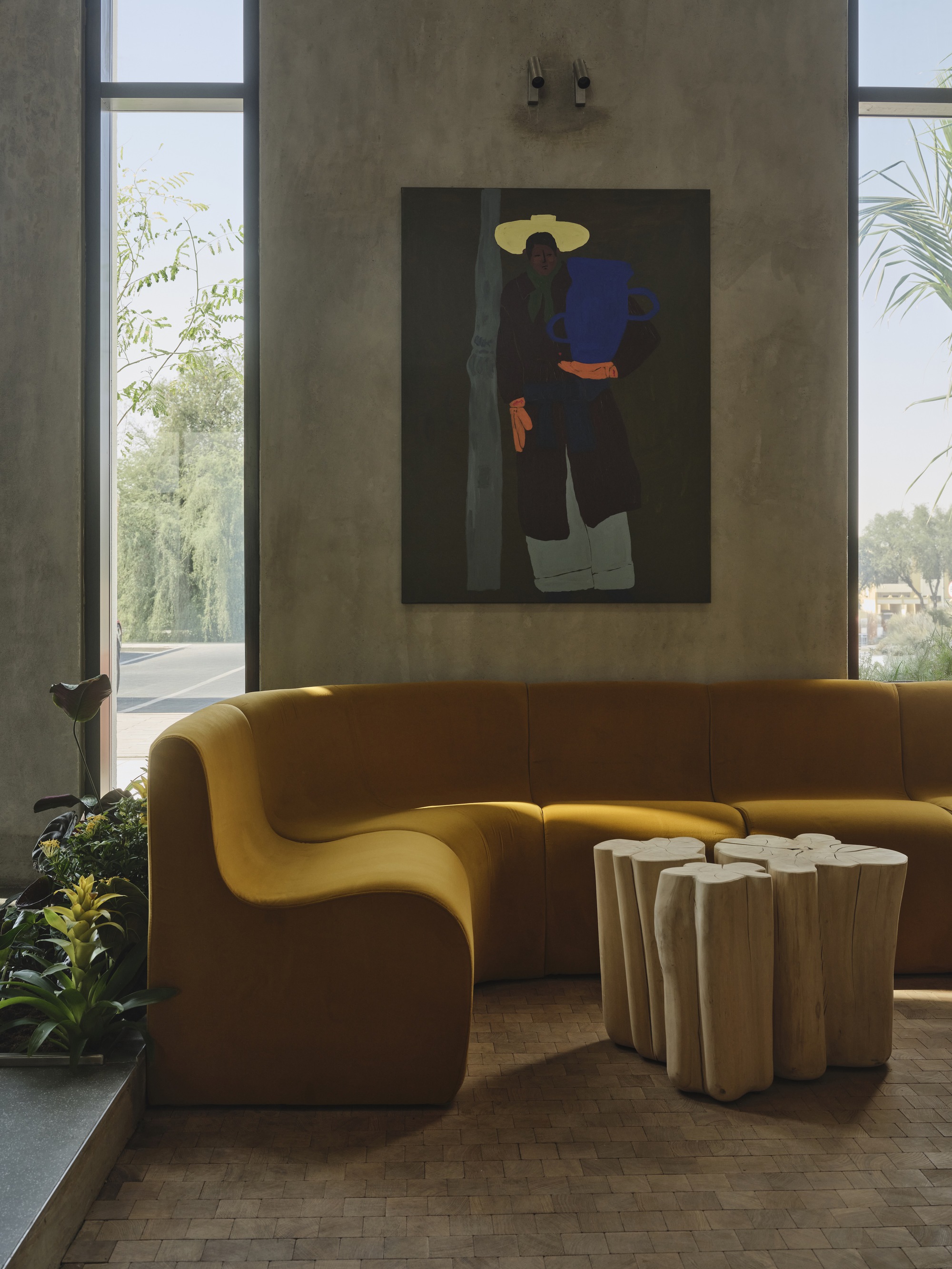 Yellow curved sofa with timber side tables beneath concrete walls and tall windows with greenery outside