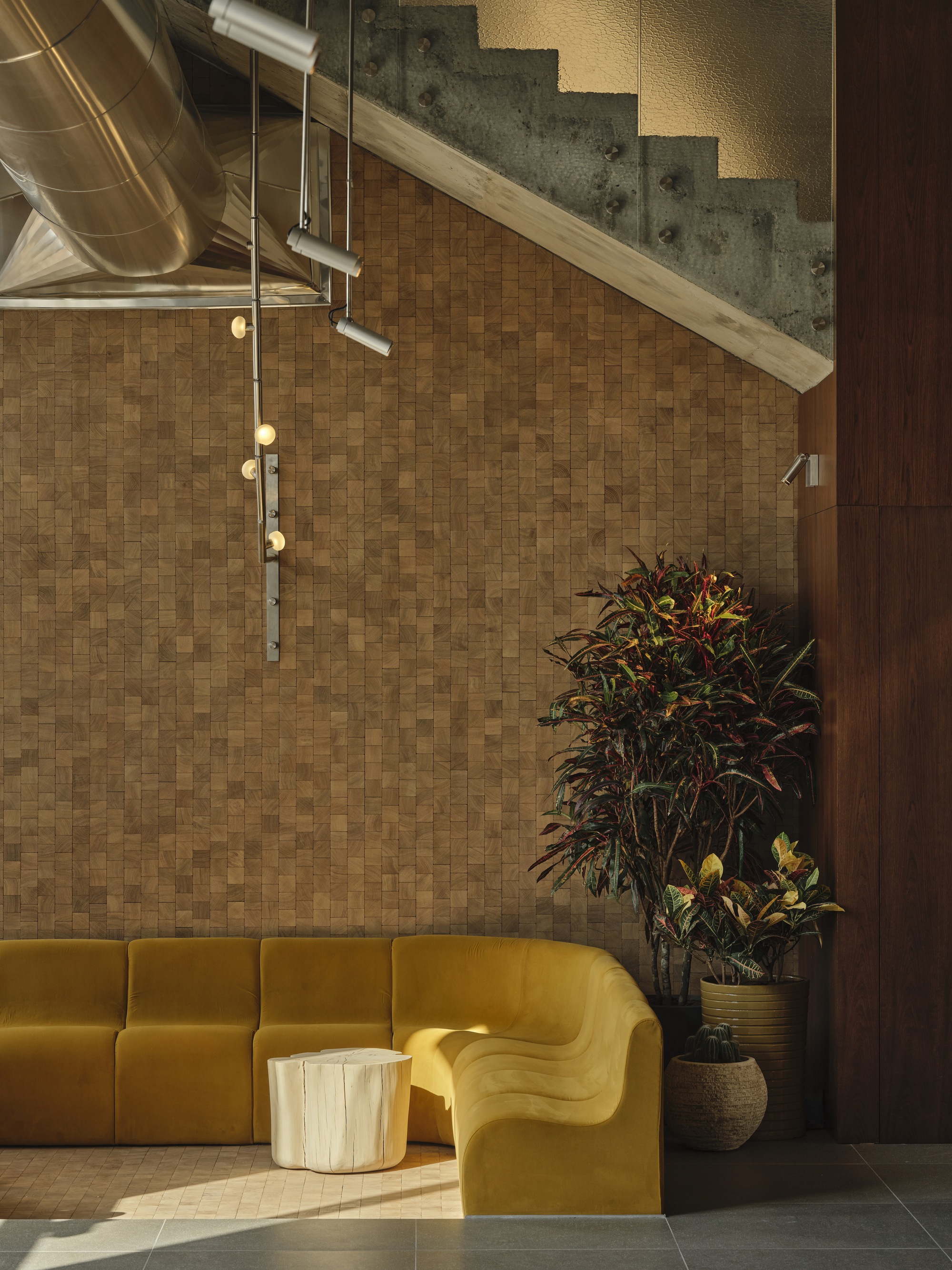 Seating area with tiled cork wall under a concrete staircase lit by pendant fixtures and ductwork