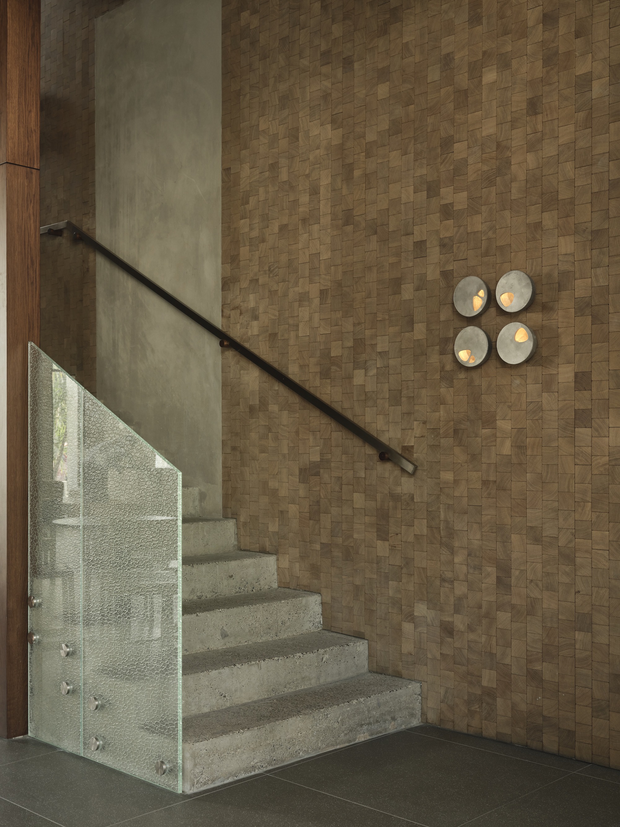 Concrete staircase with glass balustrade and metal handrail alongside a basketweave cork wall with circular sconces