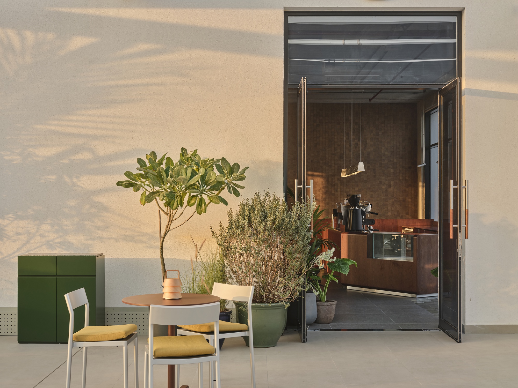 Outdoor seating area with potted plants and glass doors opening to a cafe interior in afternoon light