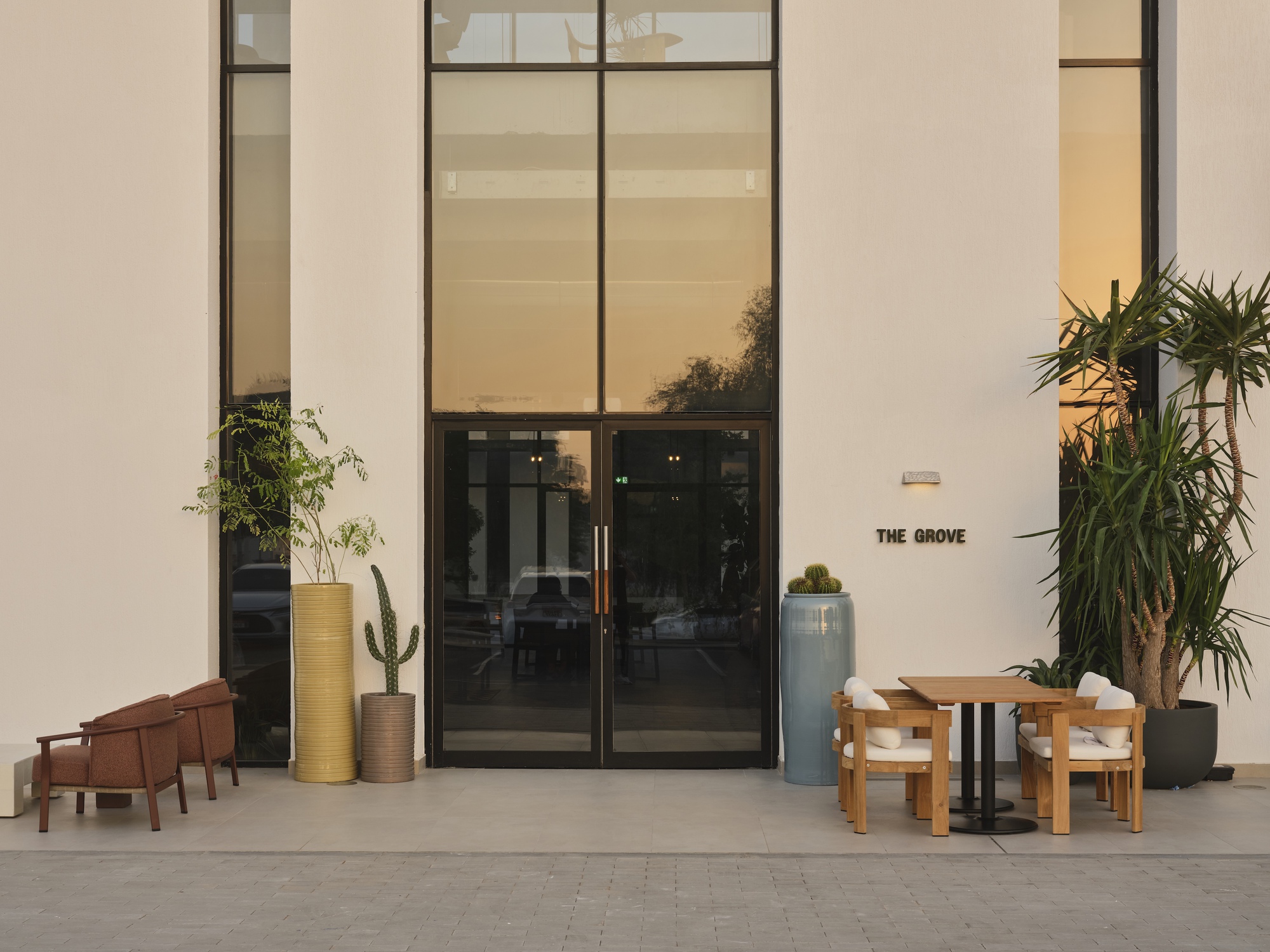 White facade with black-framed glass doors flanked by potted cacti and seating at dusk