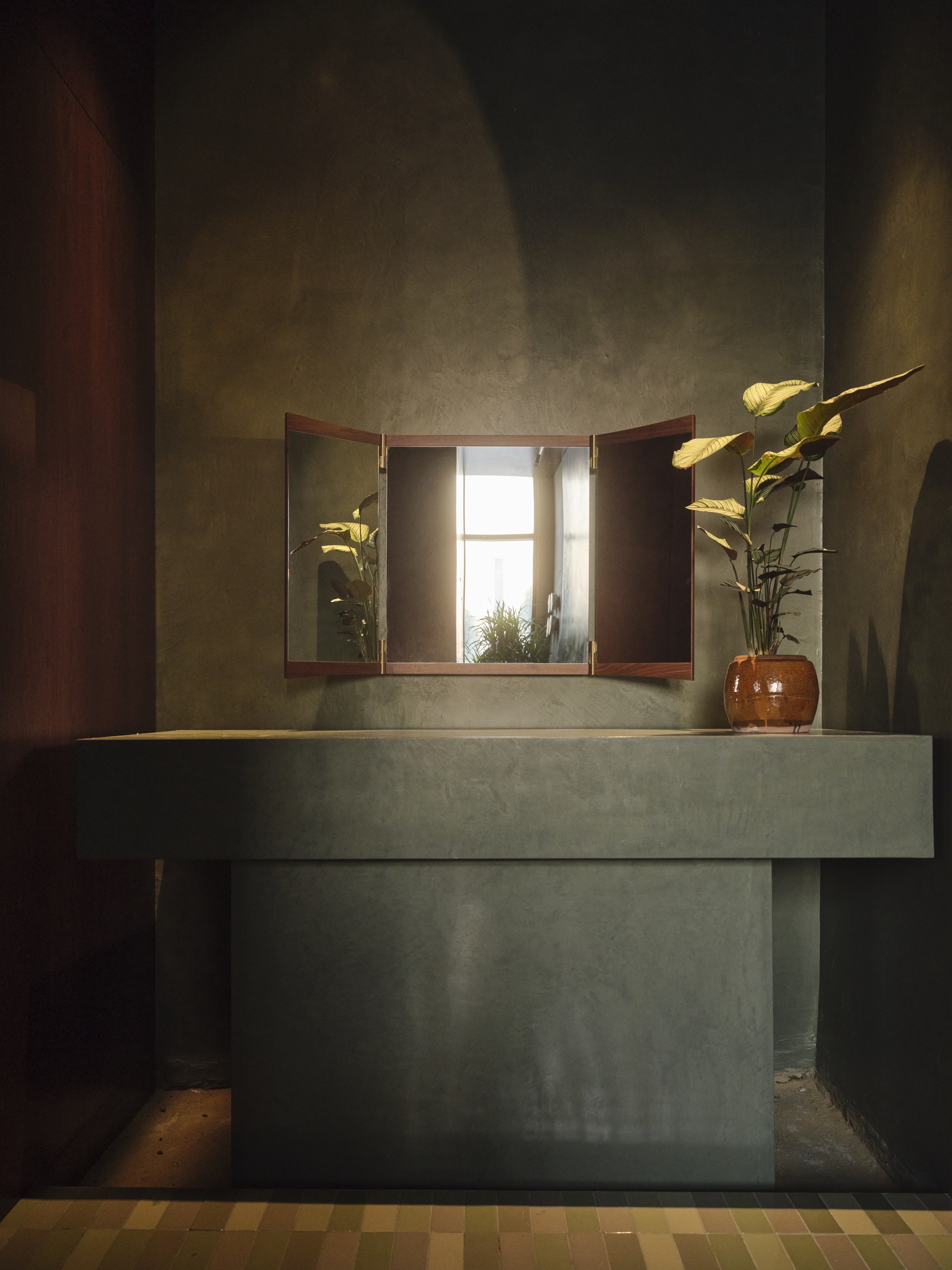 Concrete vanity unit with triptych mirror reflecting a window in a dimly lit washroom