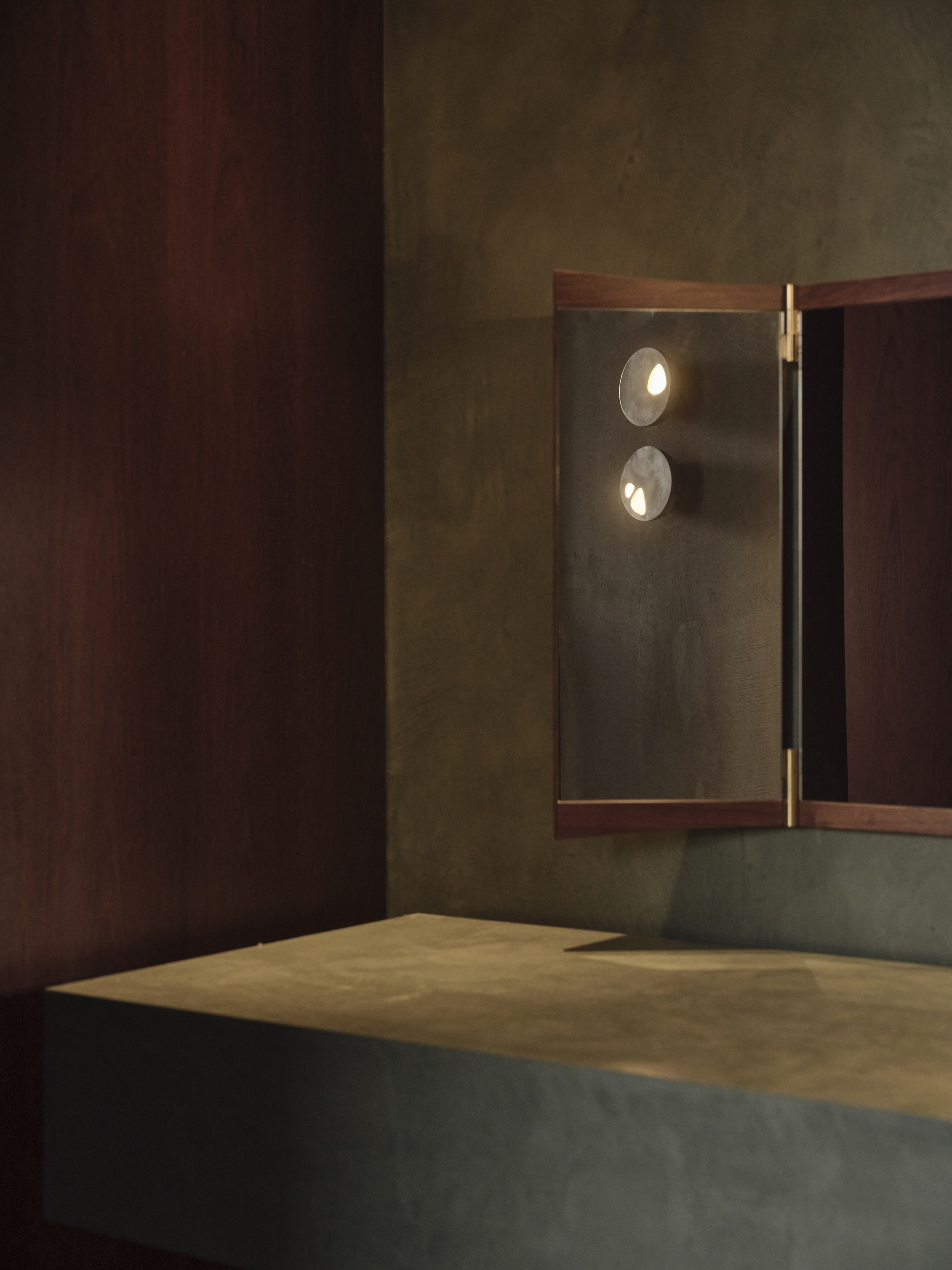 Illuminated triptych mirror panel with brass hinges set into dark timber and concrete walls