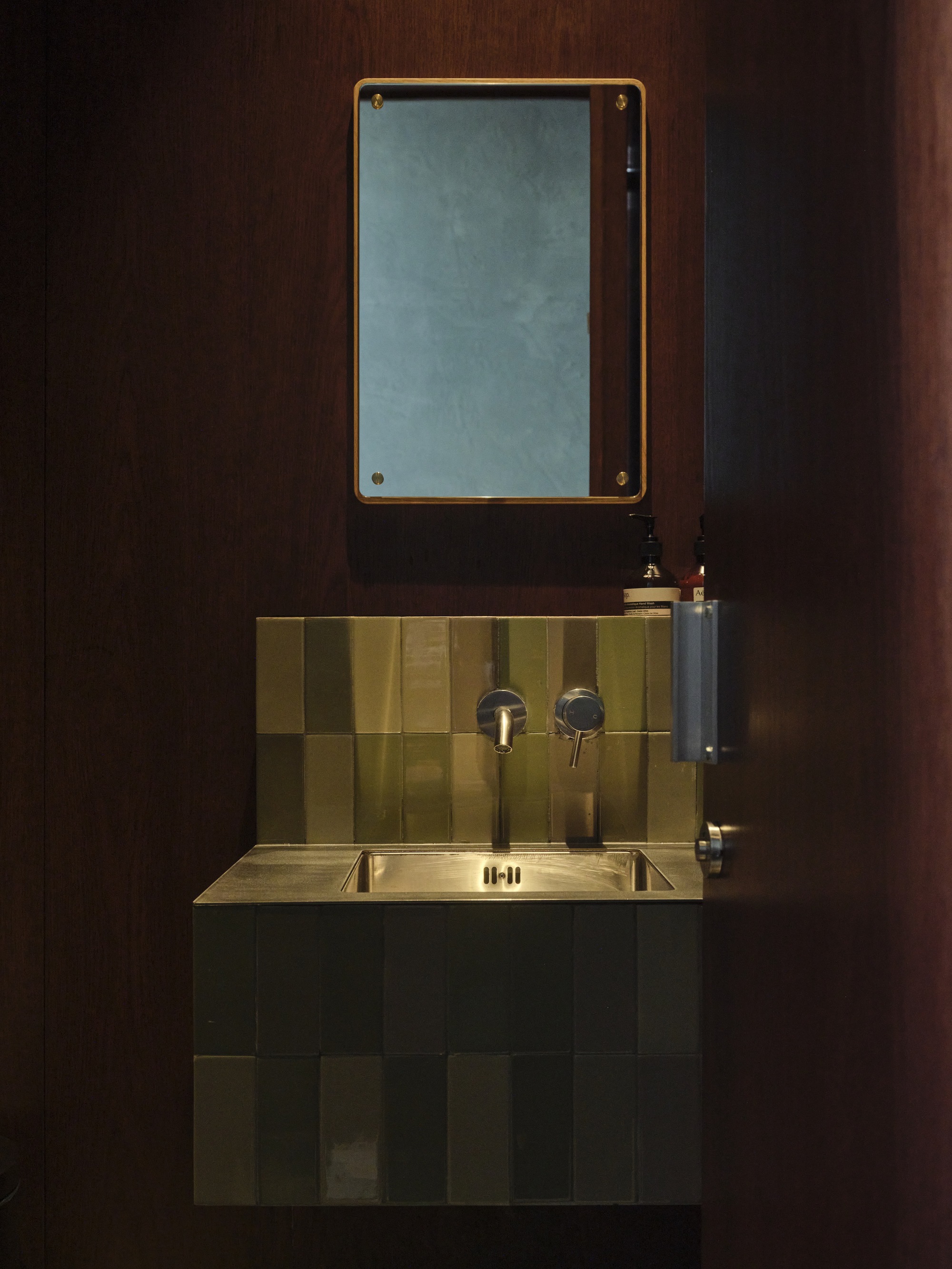 Brass-framed mirror above a checkered tile backsplash and wall-mounted sink in a dark washroom